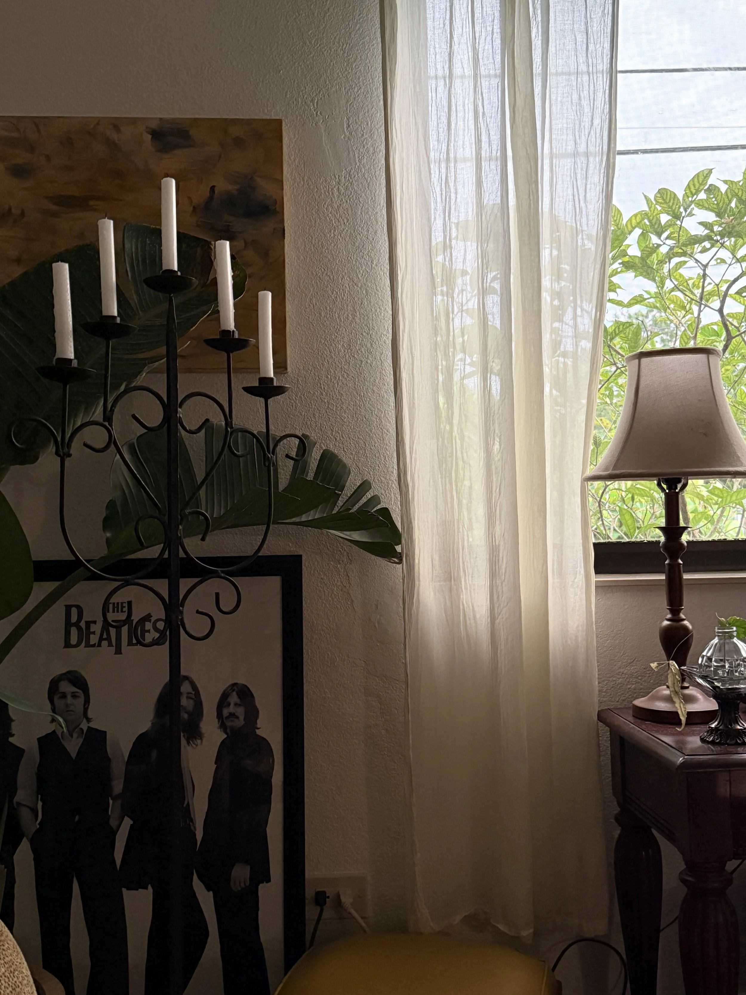 A room corner with beige curtains, a window with greenery outside, a wooden side table with a lamp and decorative items, a framed The Beatles poster, and a lit black candelabra with five white candles.