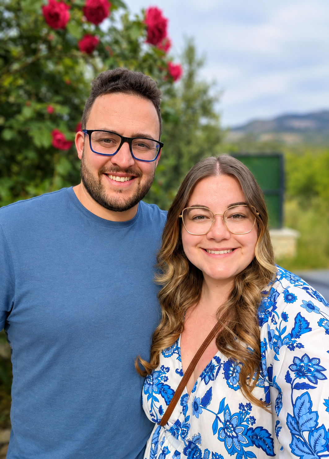 A smiling man and woman, Andy and Georgie, owners of The Heath & Acre. Andy is wearing glasses, and a blue t-shirt, and Georgie is wearing a blue and white dress. The image is a selfie taken outdoors in front of greenery and pink flowers.