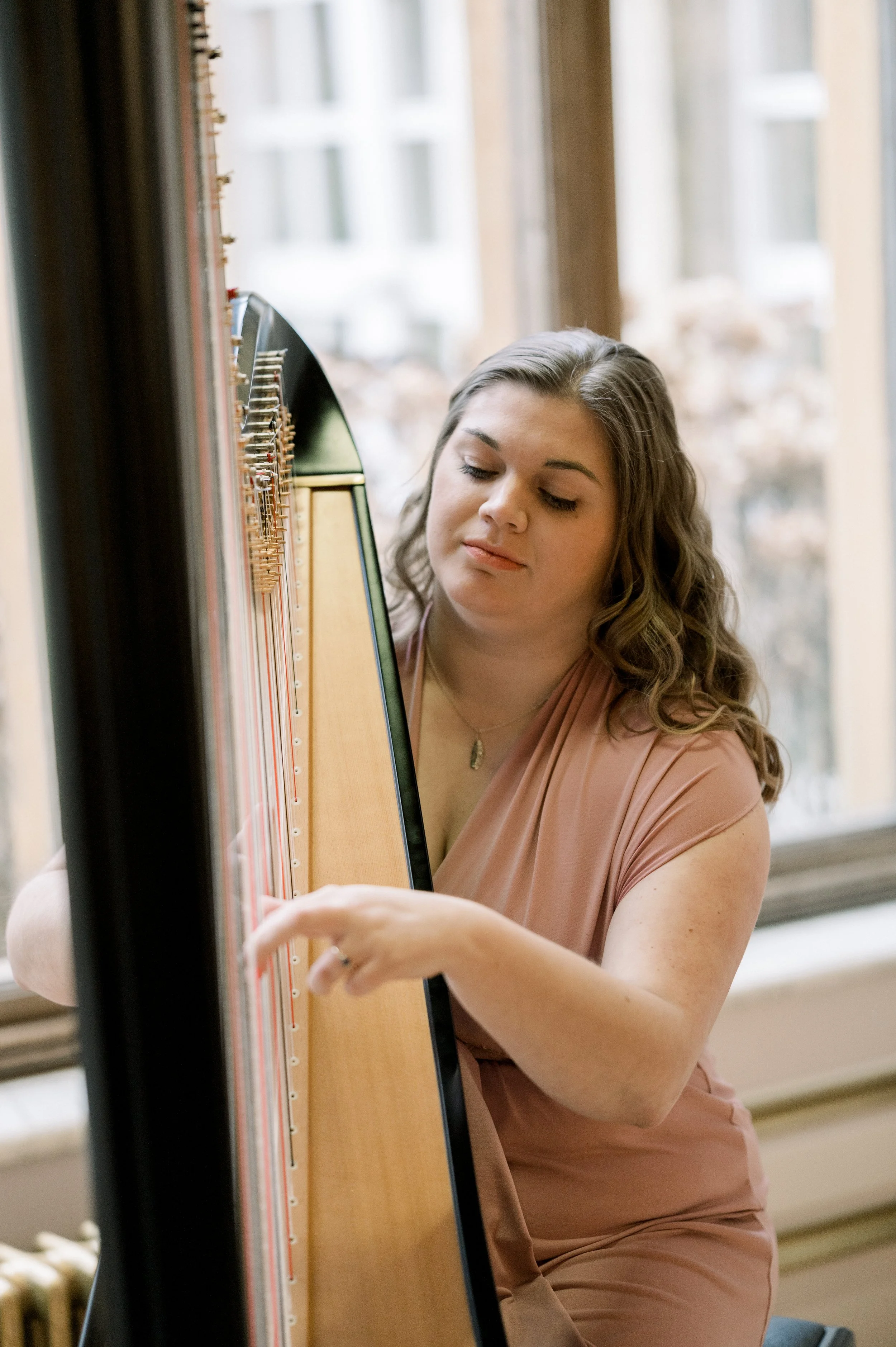A woman with long, wavy brown hair playing a harpsichord near a window with a view of buildings outside.