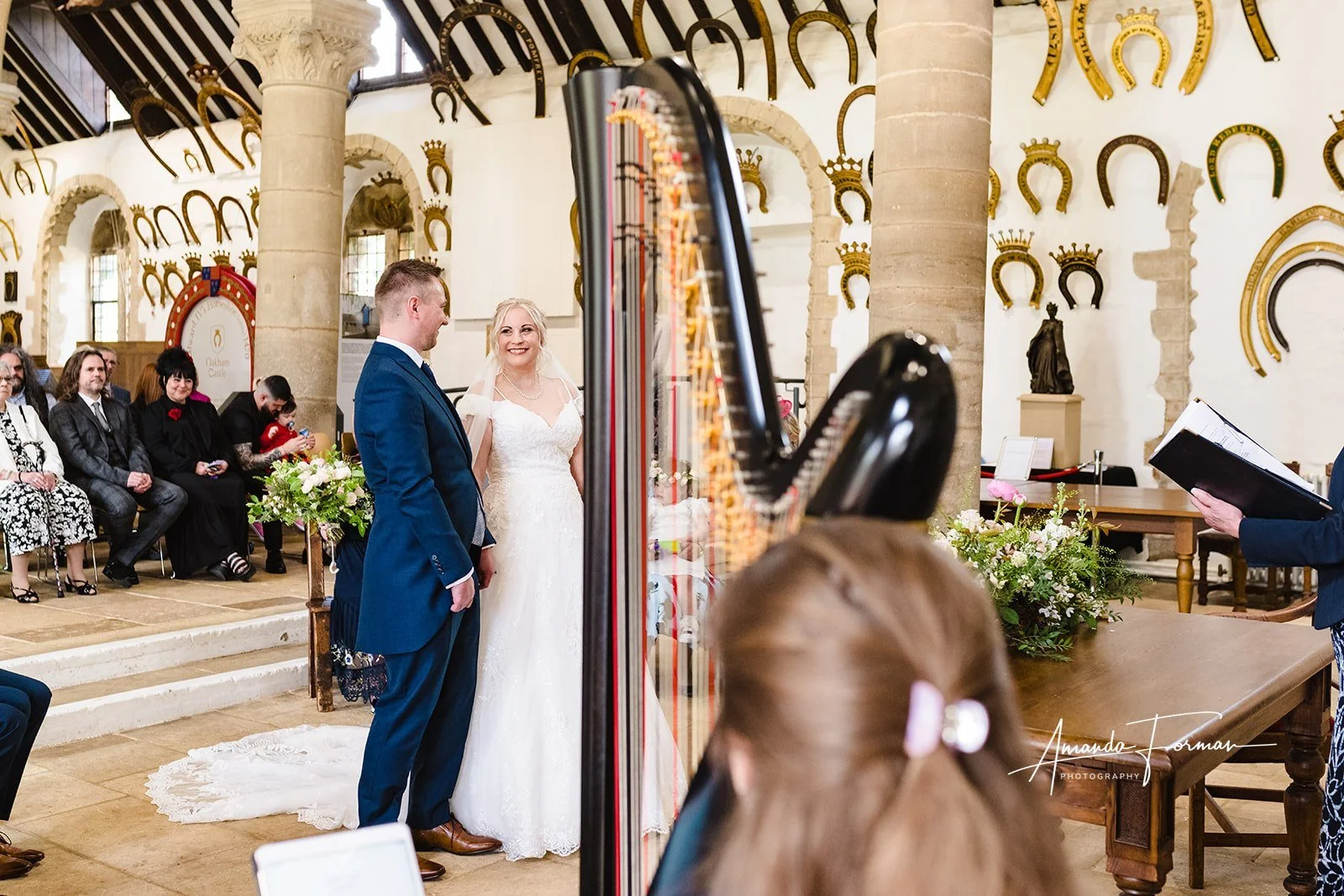 A bride and groom standing together during their wedding ceremony, surrounded by seated guests and floral arrangements in a decorated church with horseshoe-shaped wall decor.