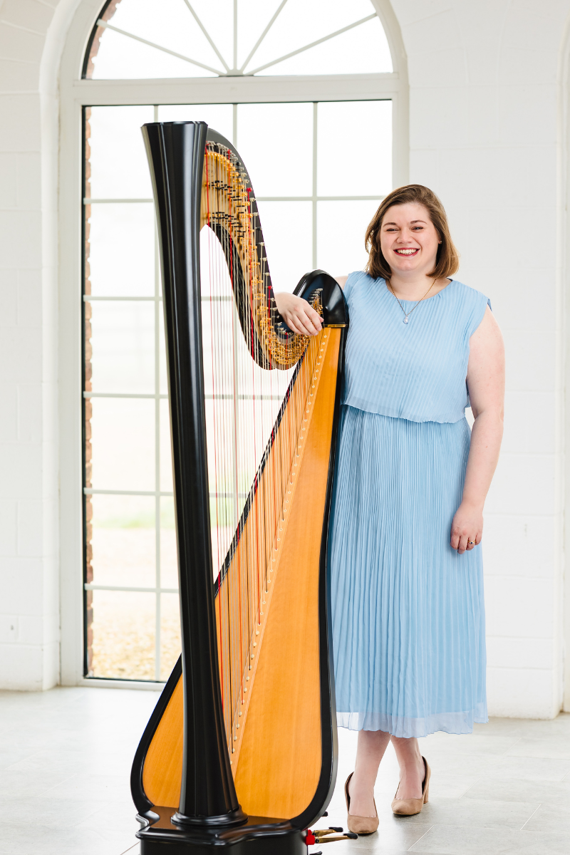 A woman in a light blue dress standing next to a harp, smiling indoors with a large arched window in the background.