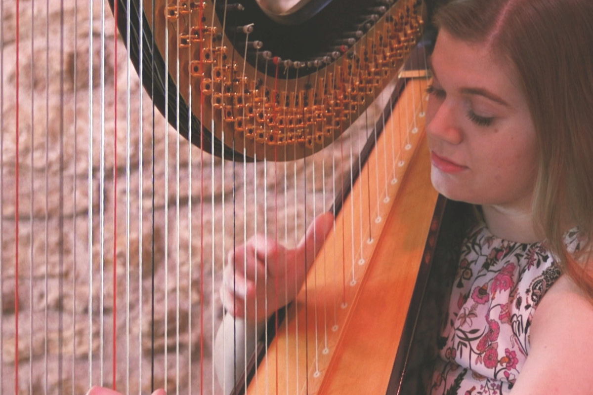 A woman with brown hair is playing a harp, with her eyes closed and a gentle smile on her face.