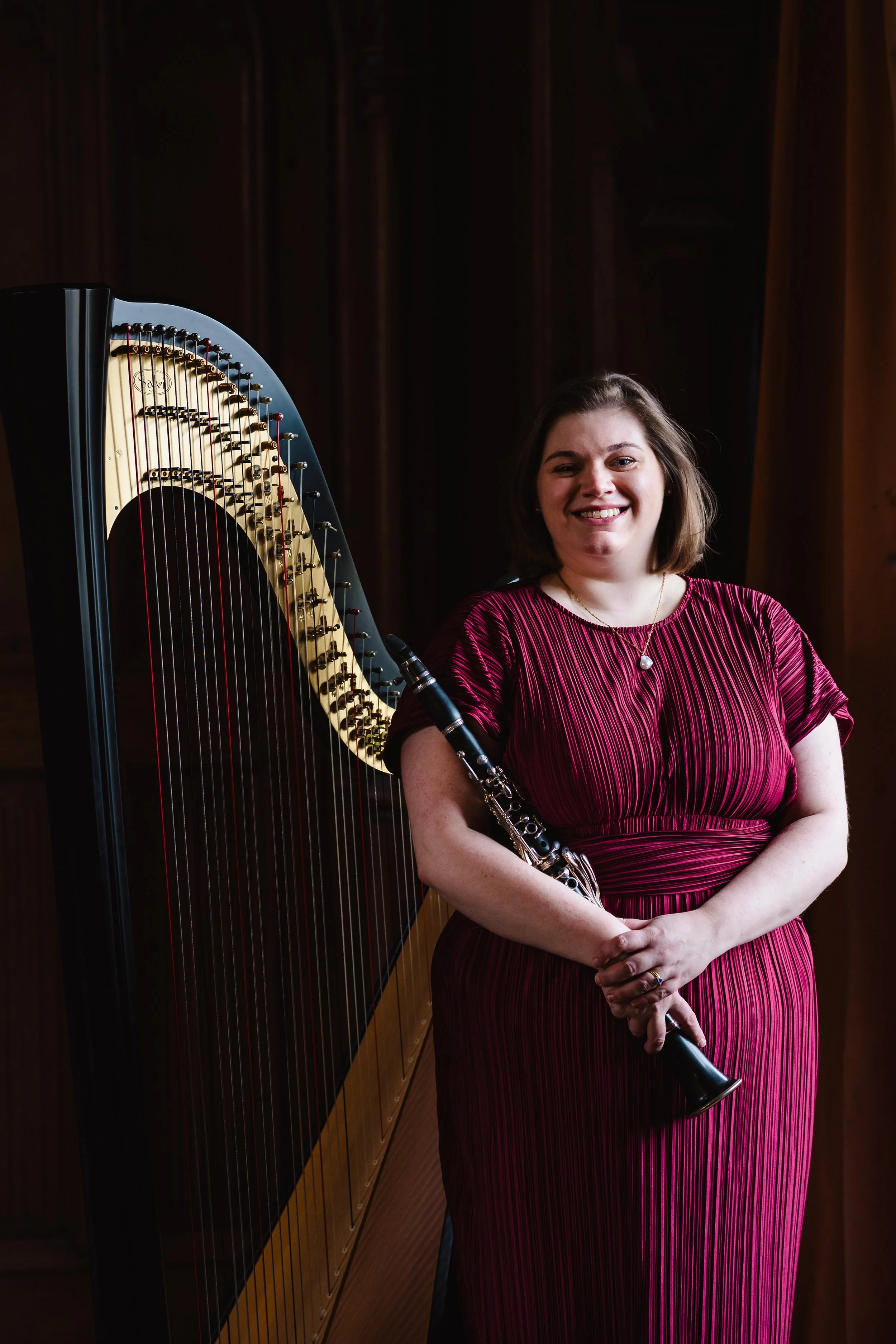 A woman in a red and black striped dress holding a clarinet, standing beside a harp in a dimly lit room with wooden walls, smiling.