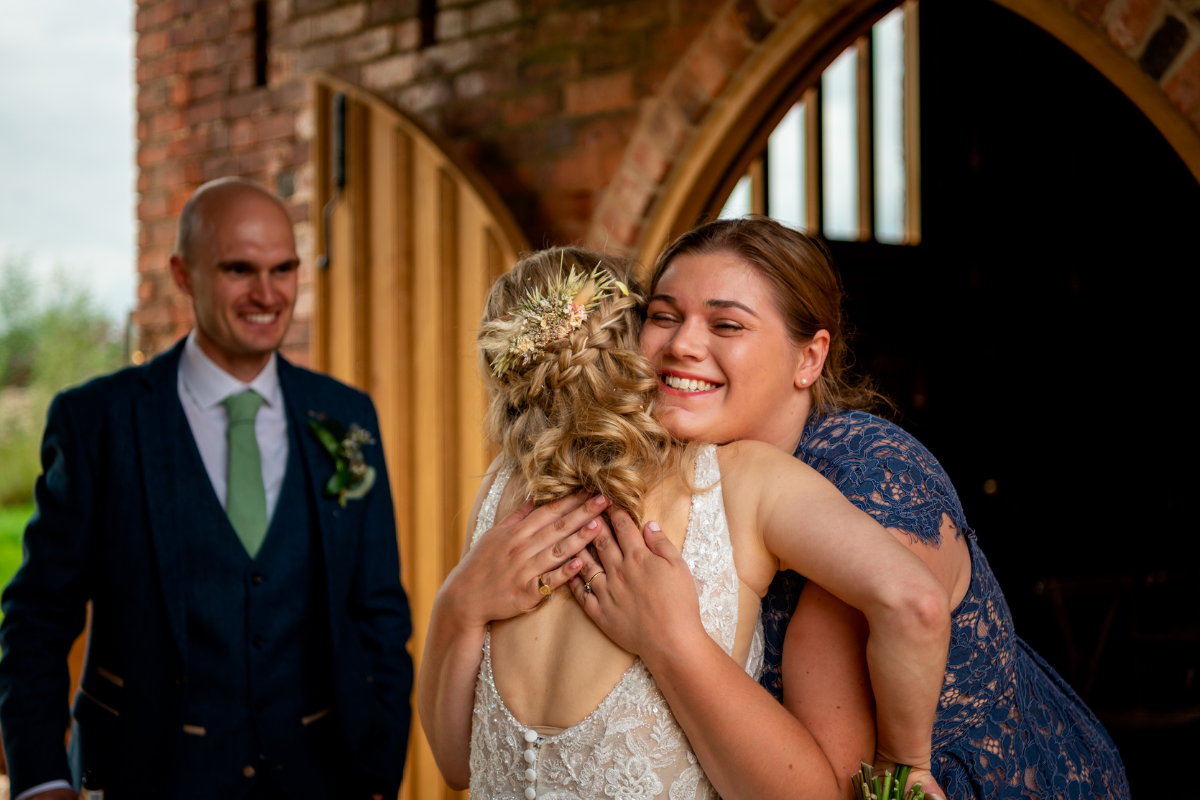 A bride hugging a woman, both smiling happily, during a wedding celebration inside a rustic venue with exposed brick and wooden doors. A man in a suit with a boutonniere is smiling in the background.