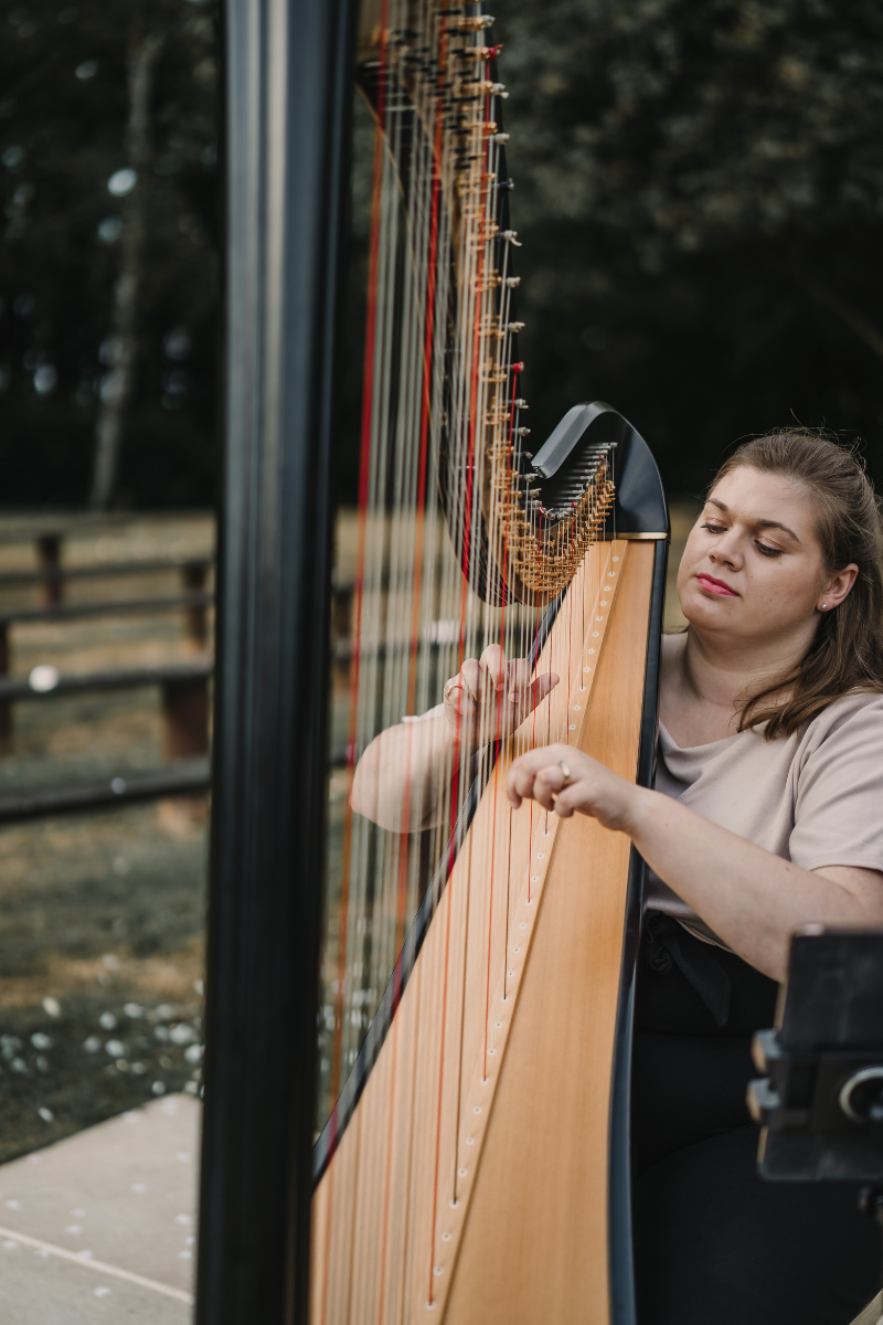 A woman playing a harp outdoors, focusing on her hands and the strings with a blurred natural background.