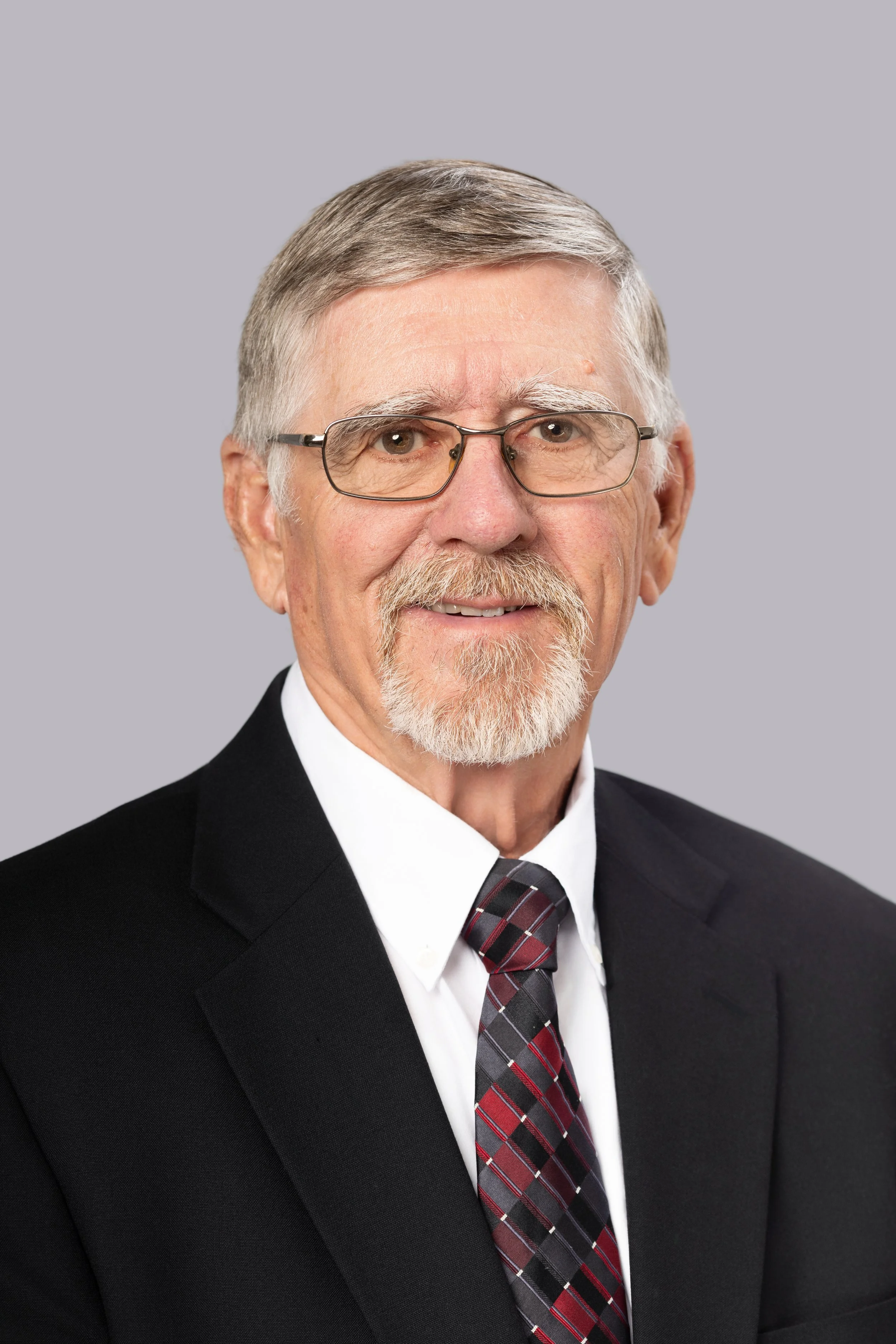 Senior man with glasses, white hair, and beard dressed in a black suit, white shirt, and a plaid tie, posing against a plain gray background.