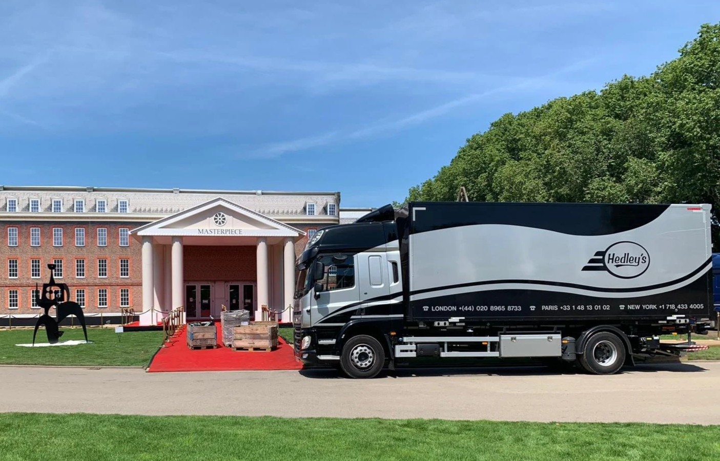 A large delivery truck with Hedley's logo parked in front of a building named 'Masterpiece,' with a red carpet, wooden pallets, and a black sculpture outside.