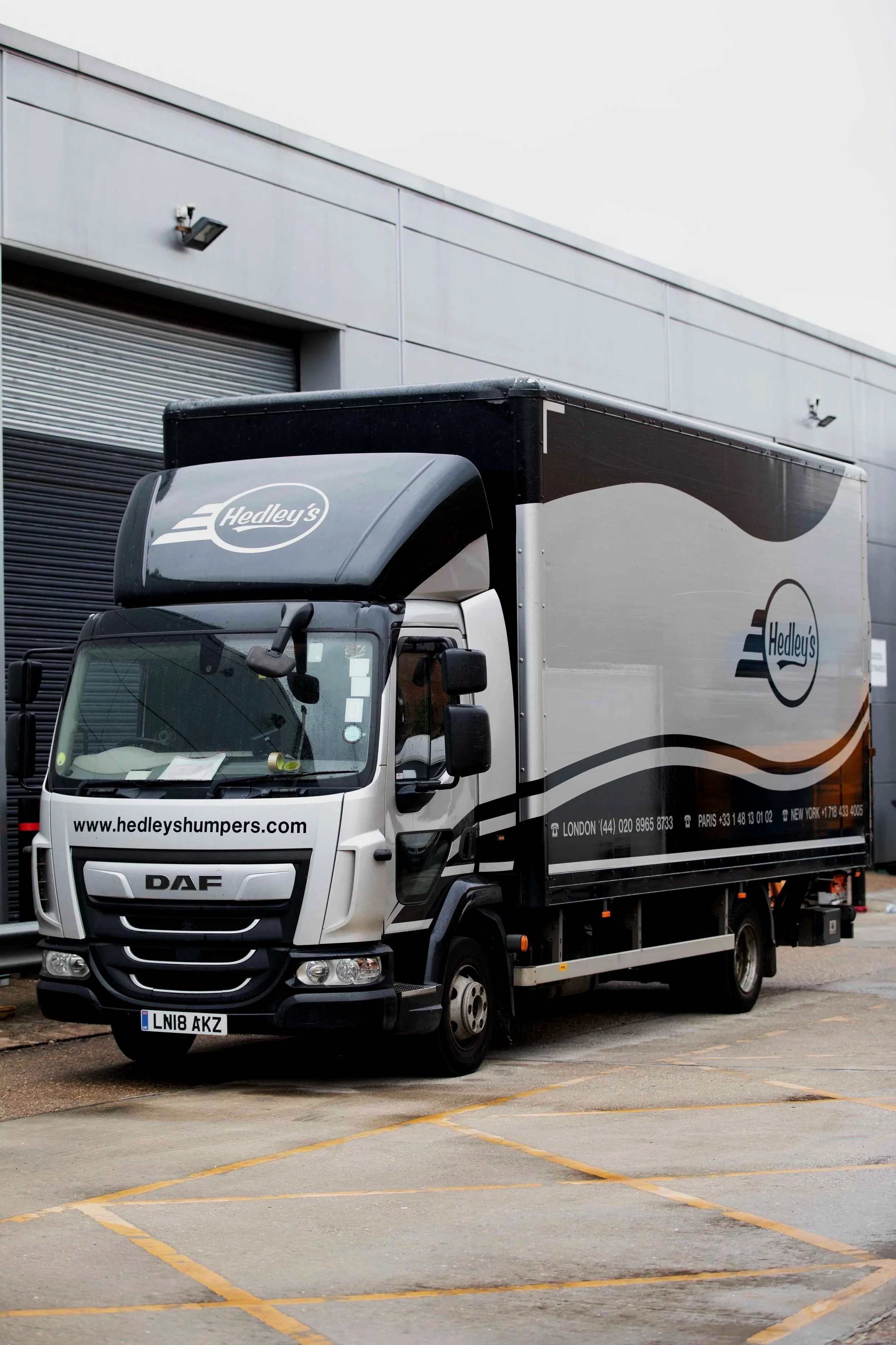 Delivery truck parked outside a building with a closed roller shutter door, displaying the Hedley's logo and website on its side and front.
