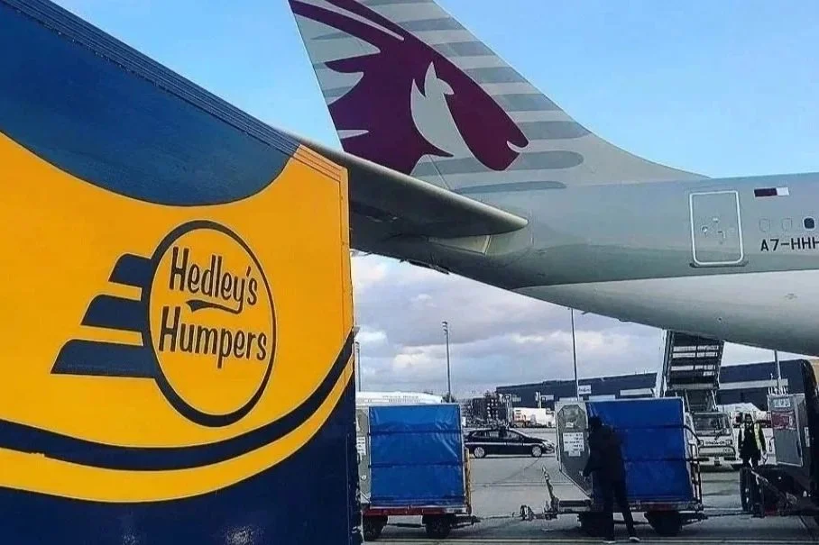 Close-up of a luggage cart with a large yellow box labeled 'Hedley's Humpers' in front of an airplane at an airport tarmac.
