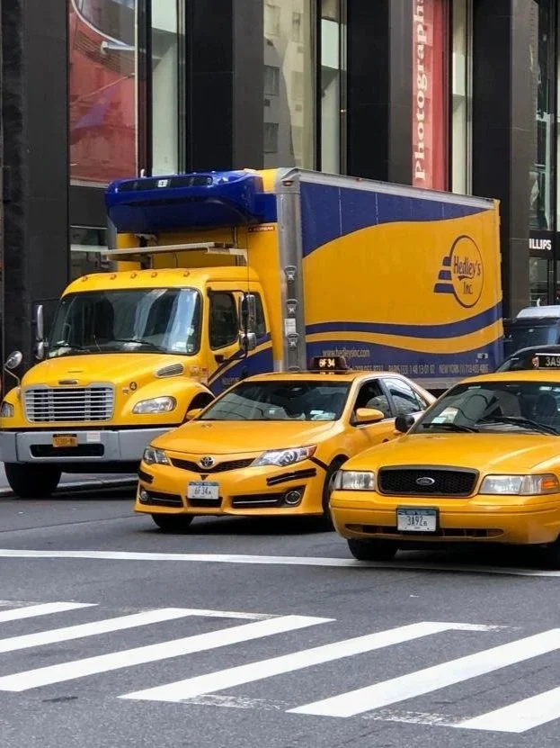 Yellow delivery truck and multiple yellow taxis parked on city street, with a crossing pedestrian crosswalk in foreground.