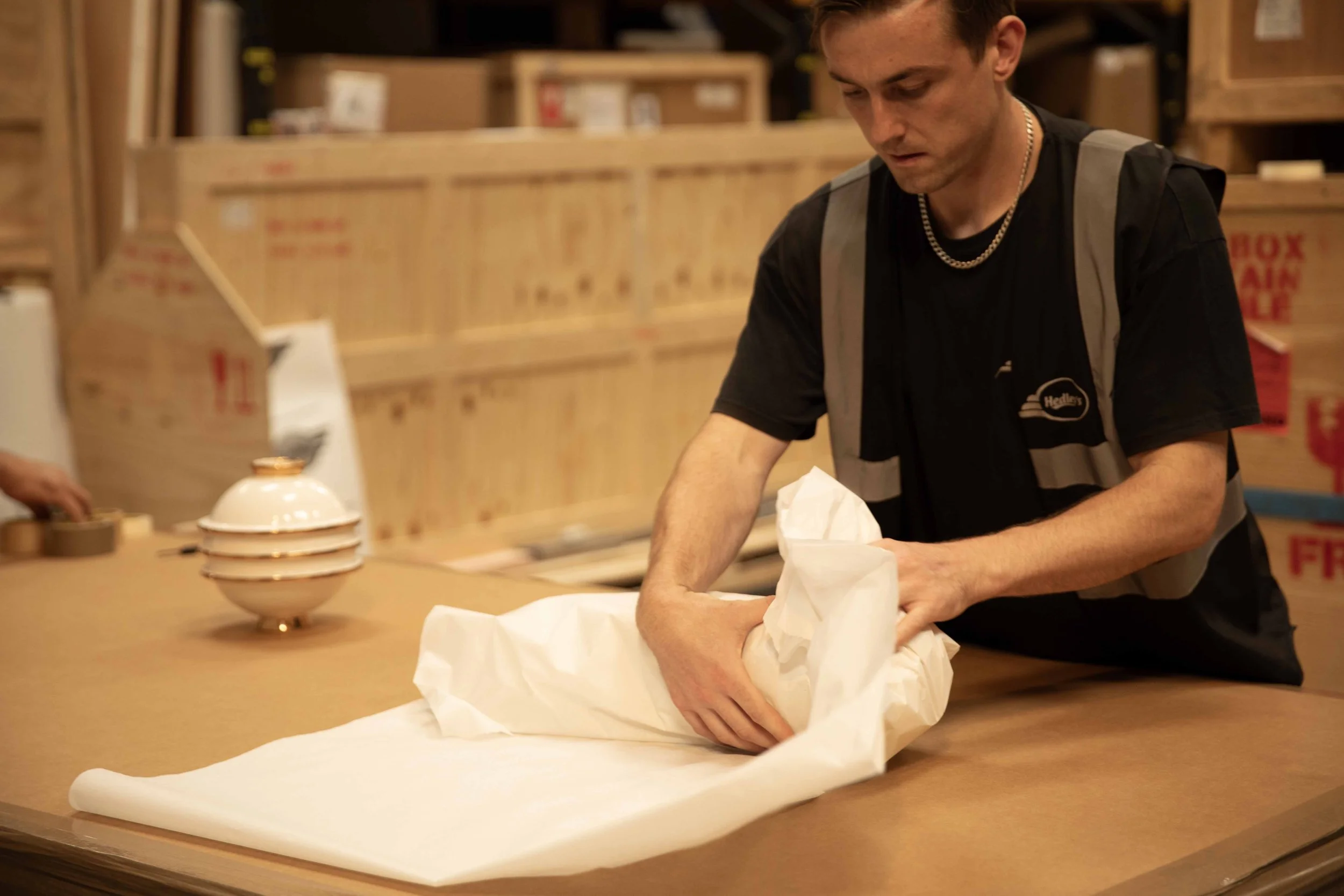 Un homme plie soigneusement du papier ou du tissu sur une table en bois dans un environnement de magasin ou d'entrepôt.