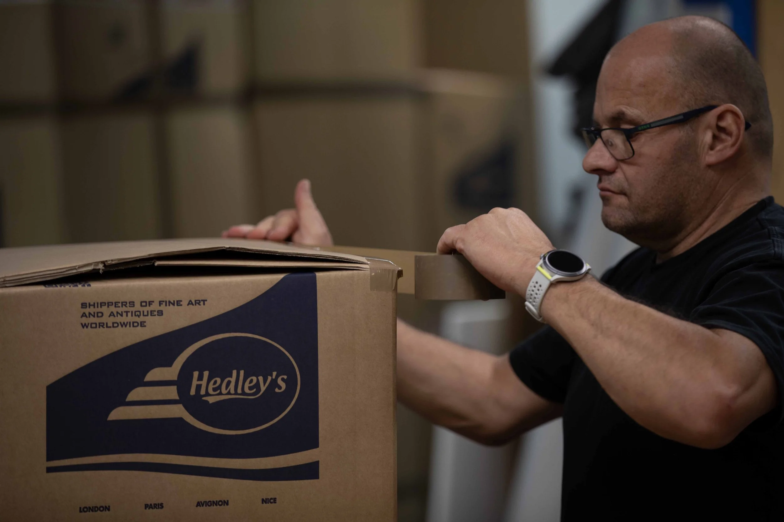 A man with glasses and a smartwatch sealing a cardboard box in a warehouse, with additional packed boxes in the background.
