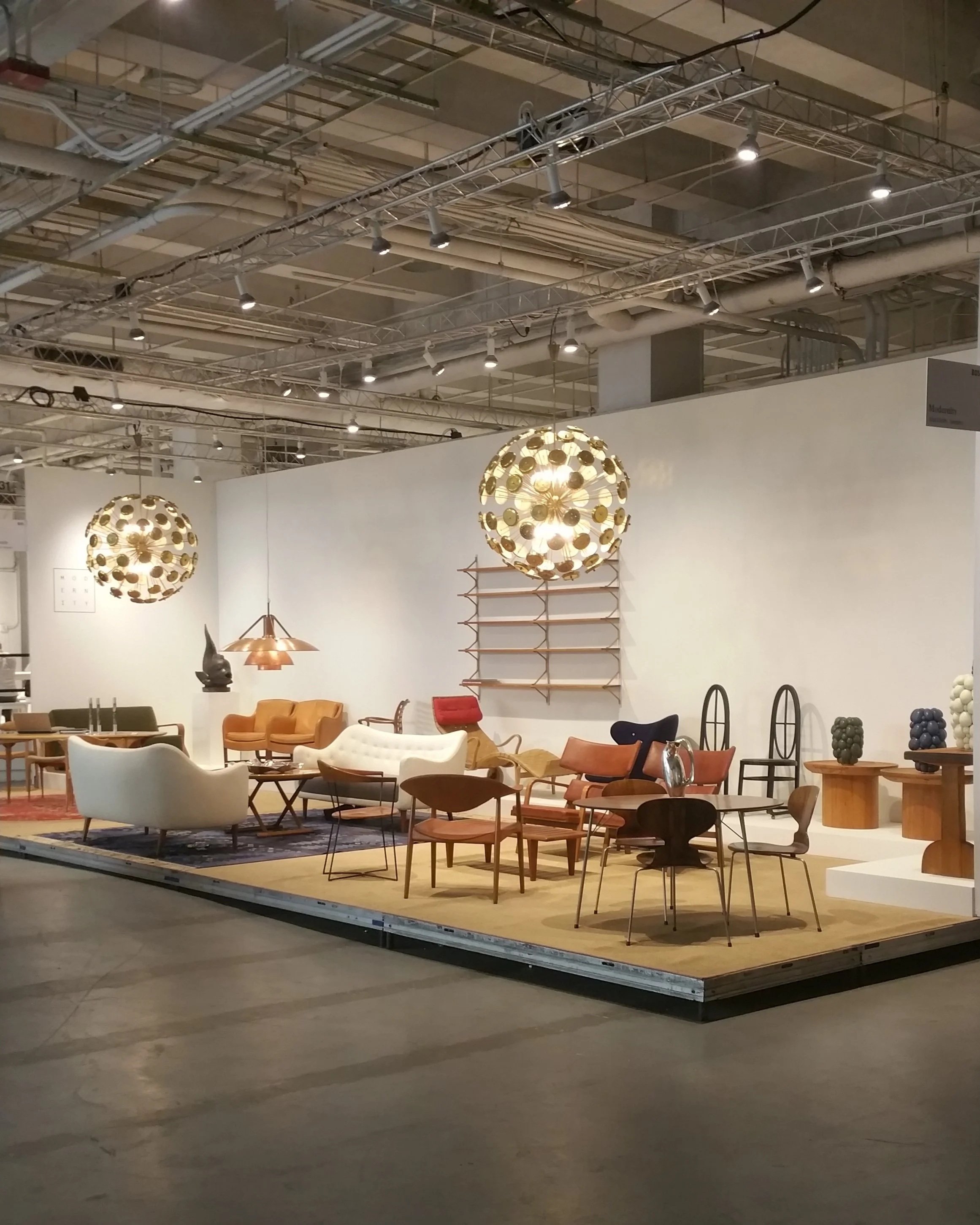 An indoor furniture display featuring various chairs, sofas, and tables on a beige rug, with decorative lighting fixtures hanging from the ceiling, and a white wall background with shelving and decorative objects.