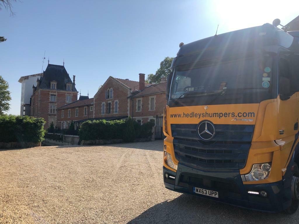 A yellow and black truck with the website 'www.hedleyshumpers.com' parked on a gravel surface in front of large historic brick buildings with pointed towers and dormer windows, under a clear blue sky.