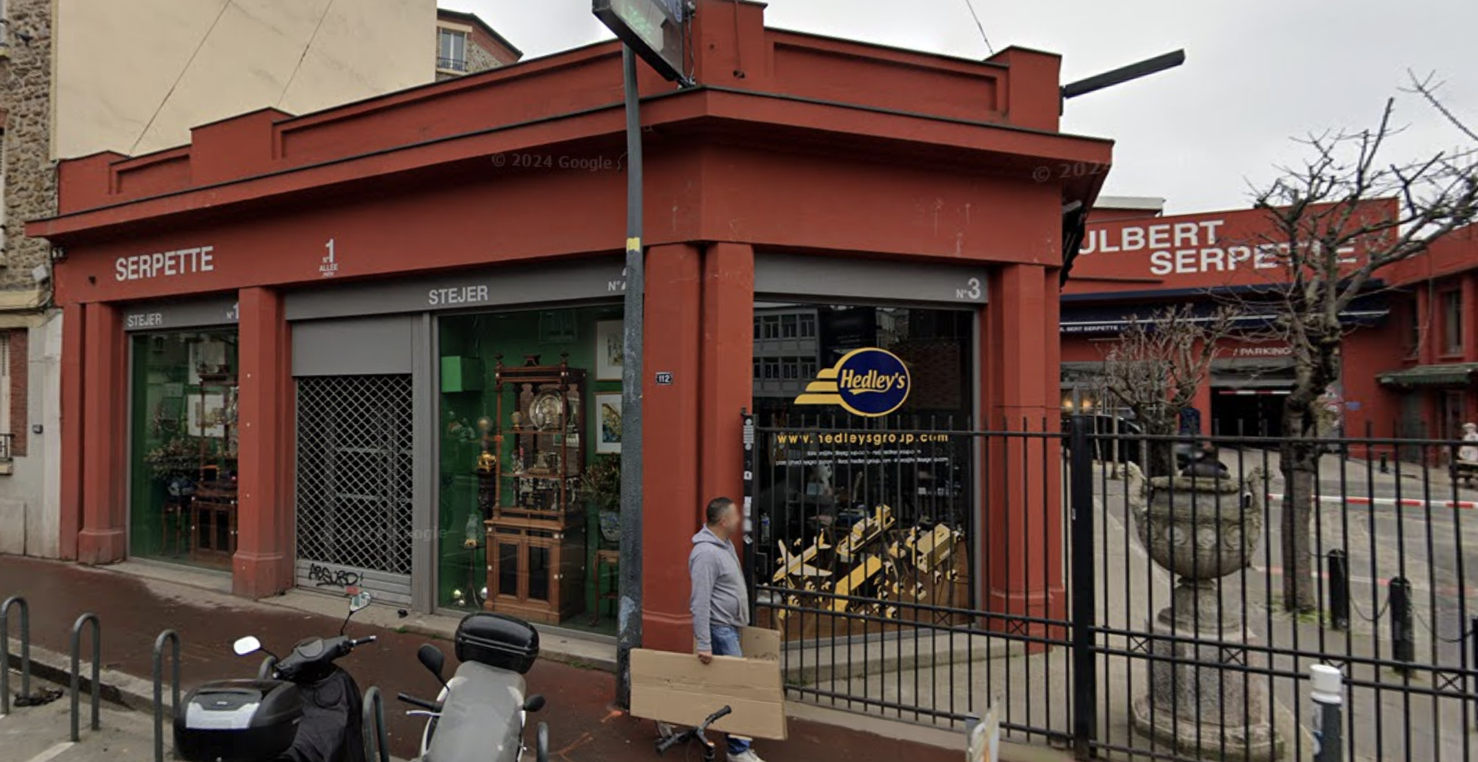 façade d'un bâtiment rouge abritant un magasin d'antiquités avec une vitrine contenant des meubles anciens, un homme avec un sac en carton à la main, abri bus en premier plan, voie de vélo et voiture à gauche, arbres sans feuilles, panneau publicitaire et grillage devant le magasin.