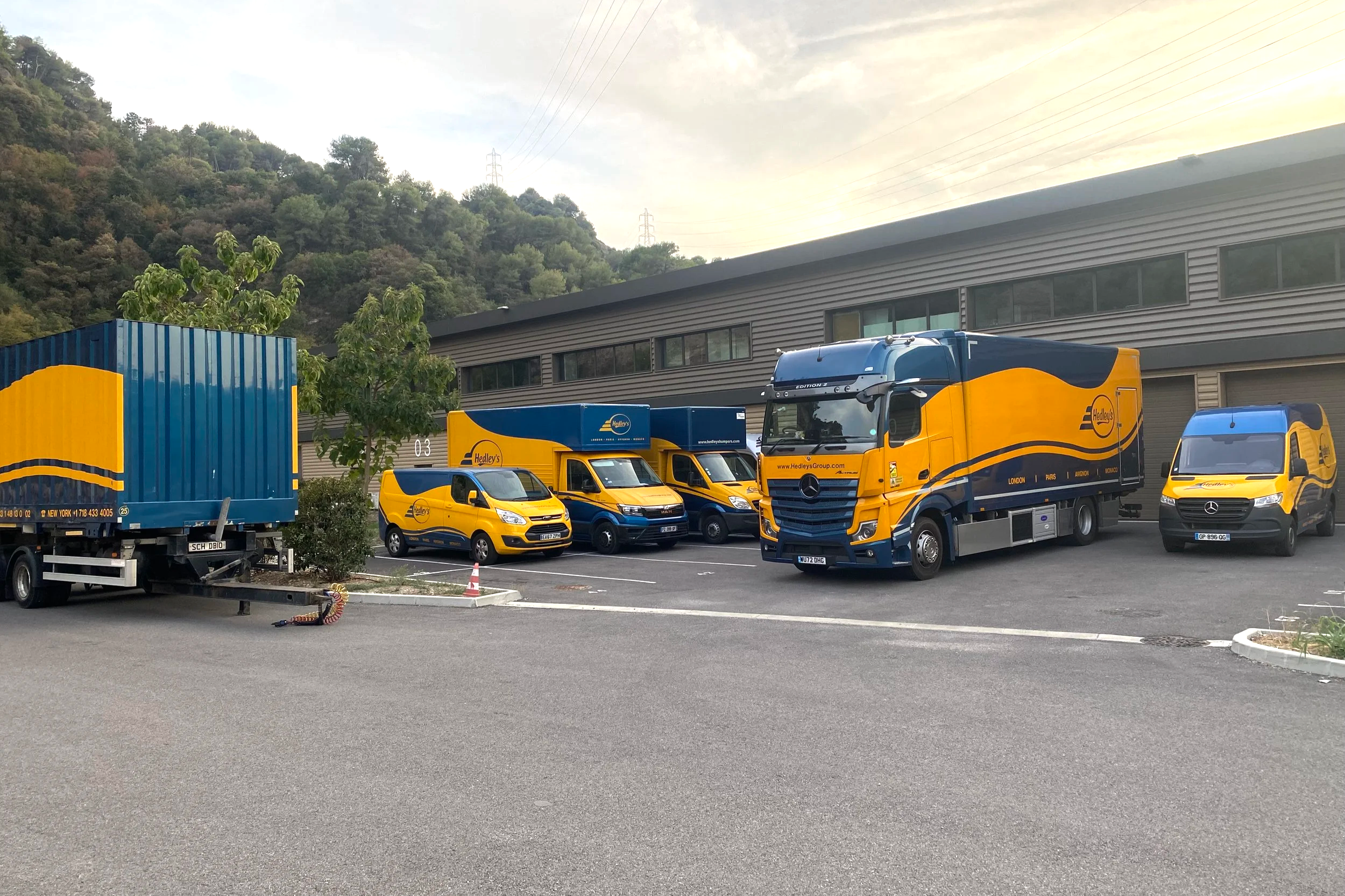 Un parking avec plusieurs véhicules de livraison de Hedley's, dont un grand camion, deux vans, et deux petites camionnettes, en face d'un bâtiment industriel et de collines verdoyantes.