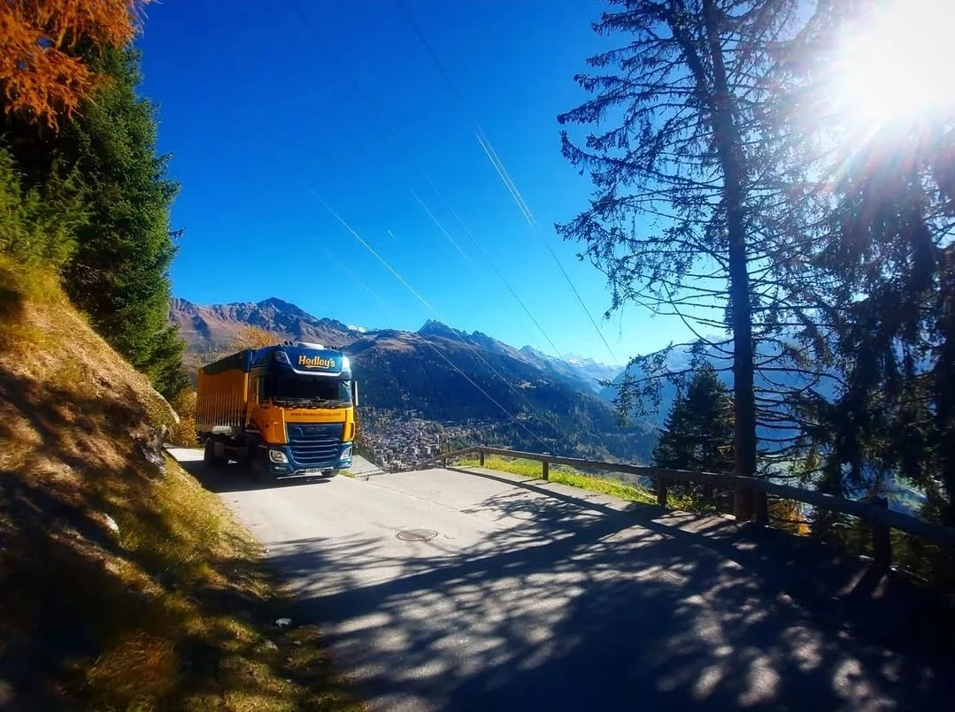 Un camion orange stationné sur une route de montagne entourée d'arbres, avec des montagnes en arrière-plan sous un ciel clair et ensoleillé.