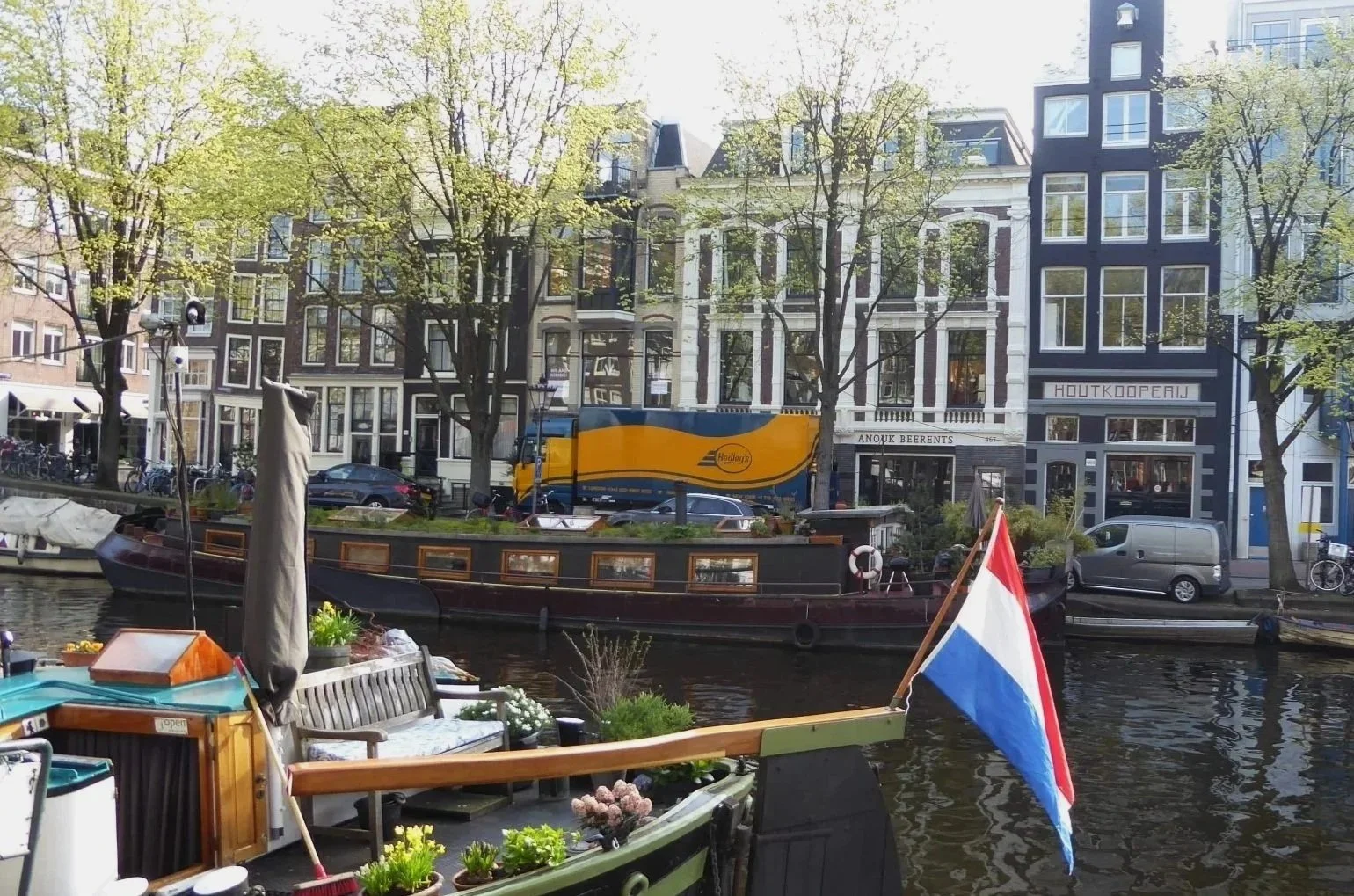 Canal avec bateau-flottant, bâtiments typiques d'Amsterdam, arbres, véhicules en stationnement, personne, commerce riverain, ciel clair.