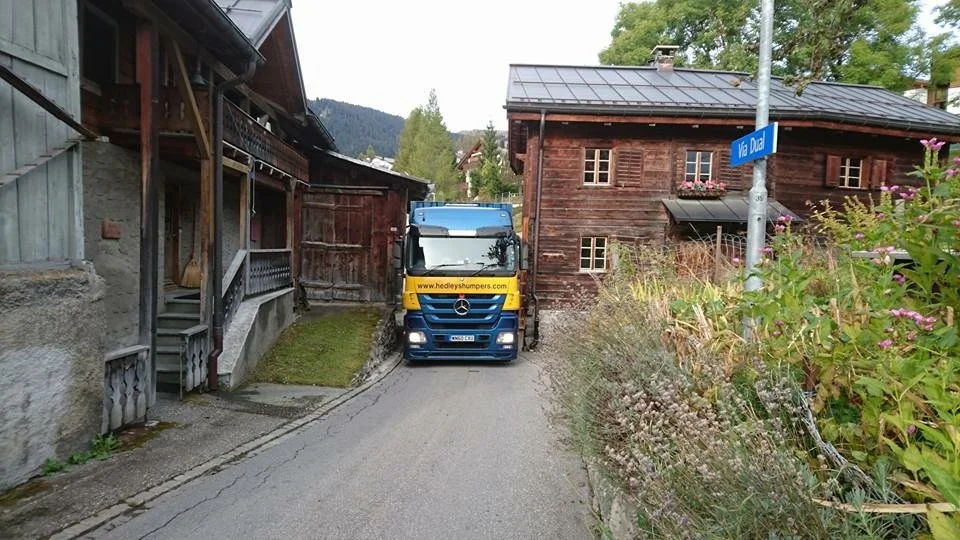 Un camion Mercedes-Benz jaune et bleu bloquant une petite route étroite dans un village européen, entouré de maisons en bois et de végétation.