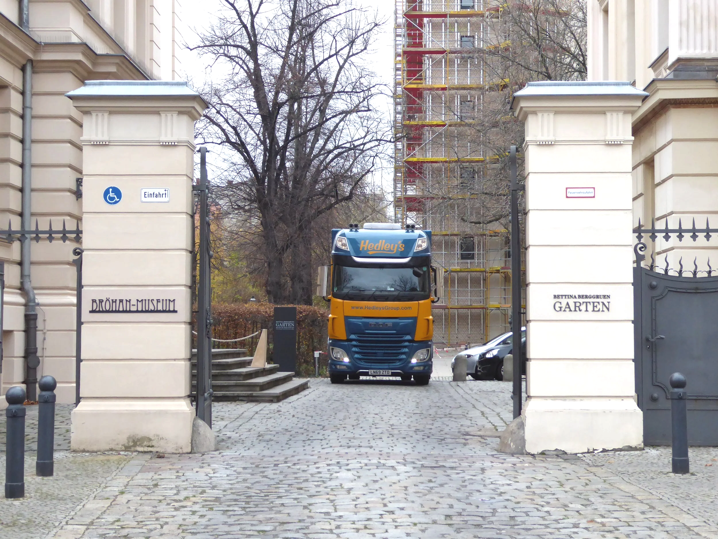 Une entrée de musée avec deux colonnes en pierre, une porte grillagée, un camion de déménagement jaune et bleu avec le nom Hedley's en haut et le site web www.Hedley'sGroup.com sur le devant, et des voitures garées à l'arrière, un arbre sans feuilles et un bâtiment en construction