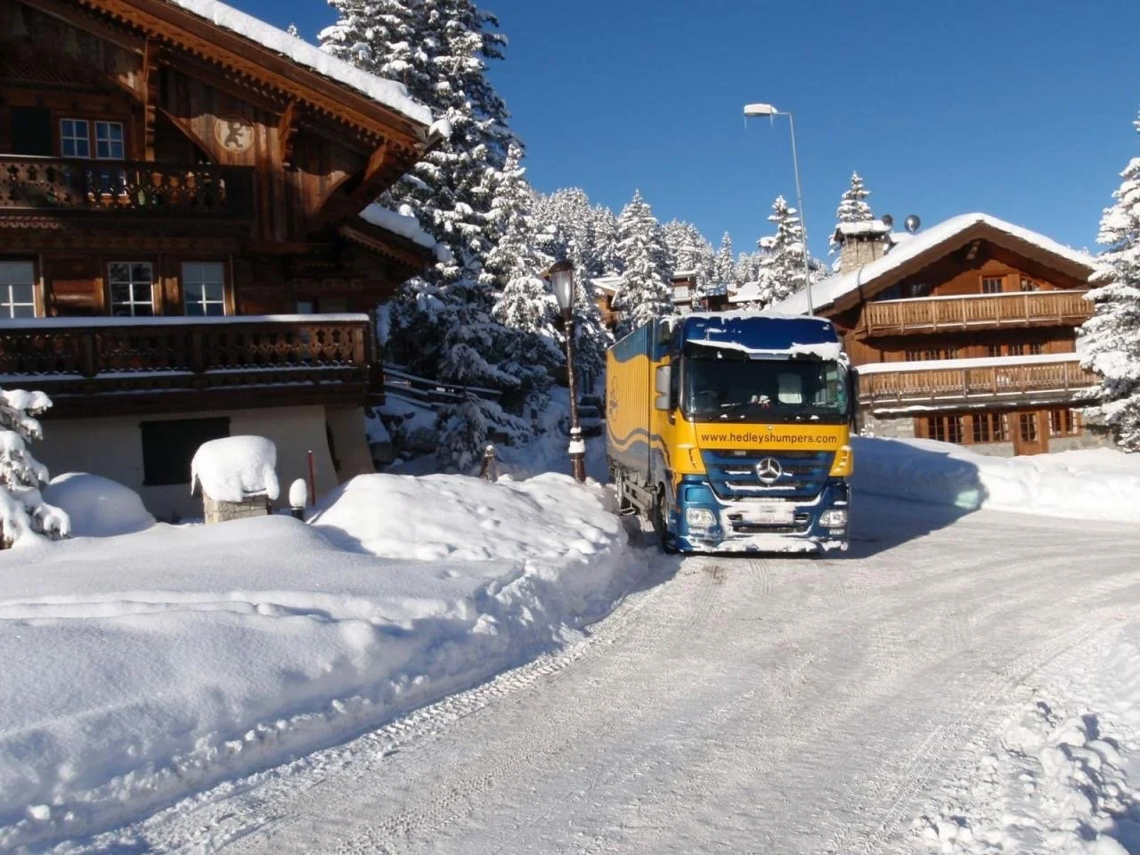 Un camion Mercedes-Benz jaune et bleu garé sur une route enneigée dans un paysage de montagne avec des chalets en bois, des sapins et un ciel bleu clair.