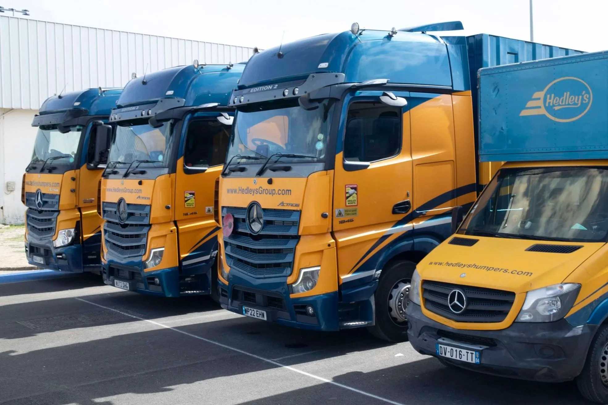 Line of three large yellow and blue trucks parked in a lot, with a smaller yellow delivery van parked in front of them. All vehicles display branding for Hedley's Group and Mercedes-Benz logos.