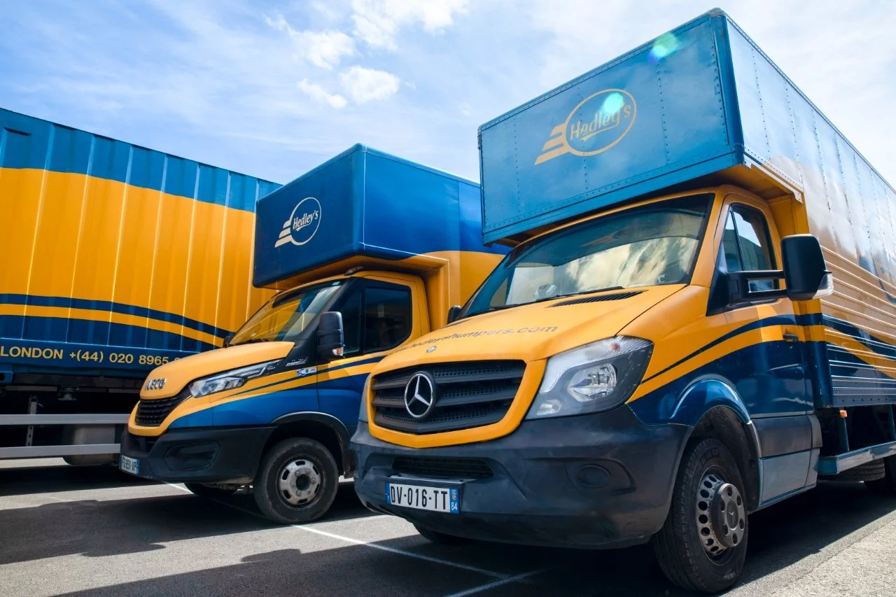 Two delivery trucks with Hedley's logo parked in a lot, painted yellow and blue, with a blue sky above.