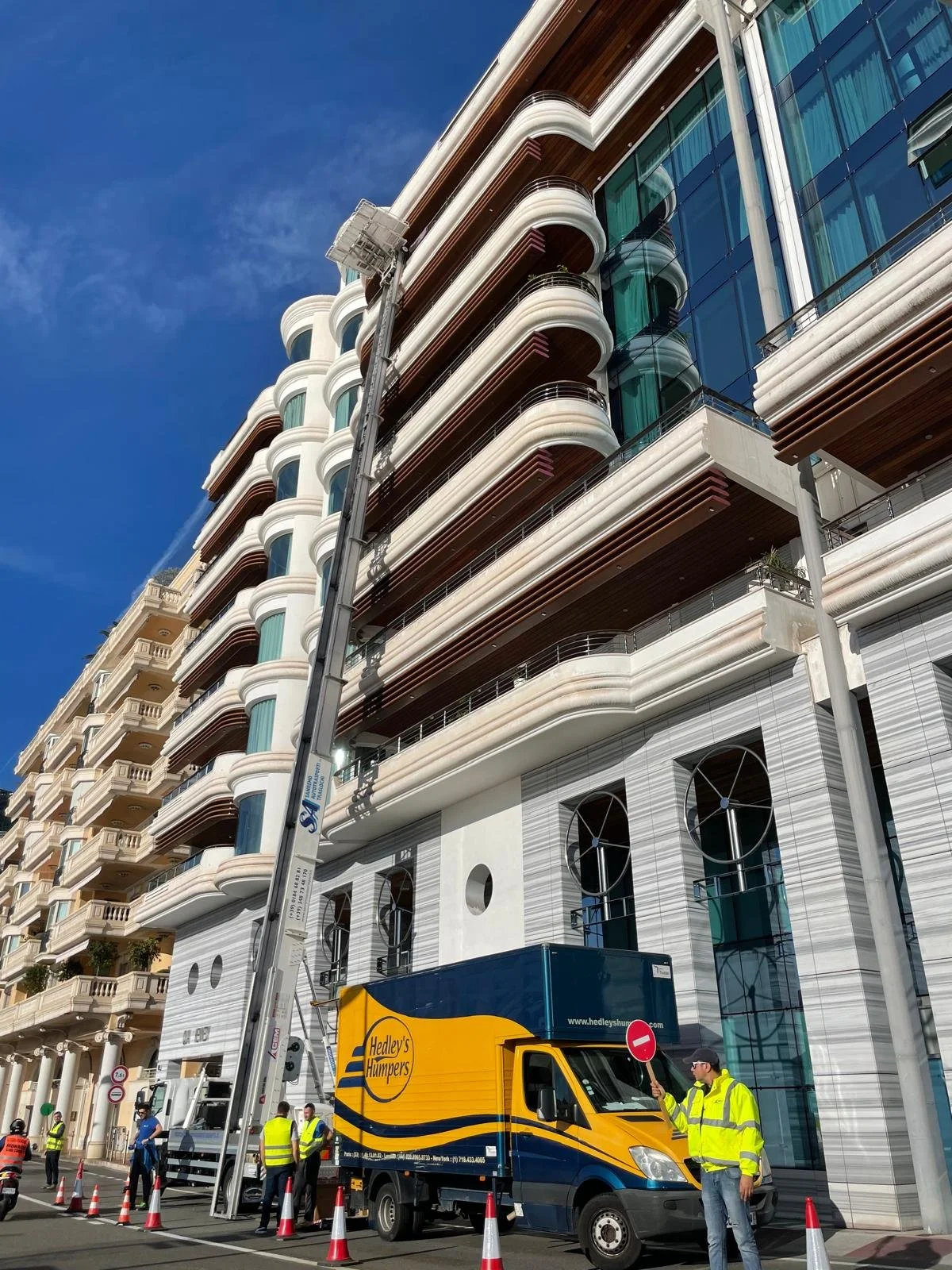 Construction crew in high-visibility jackets and hard hats working on a tall modern building with glass balconies, with a crane and a Hedley's Humper truck on the street in front.