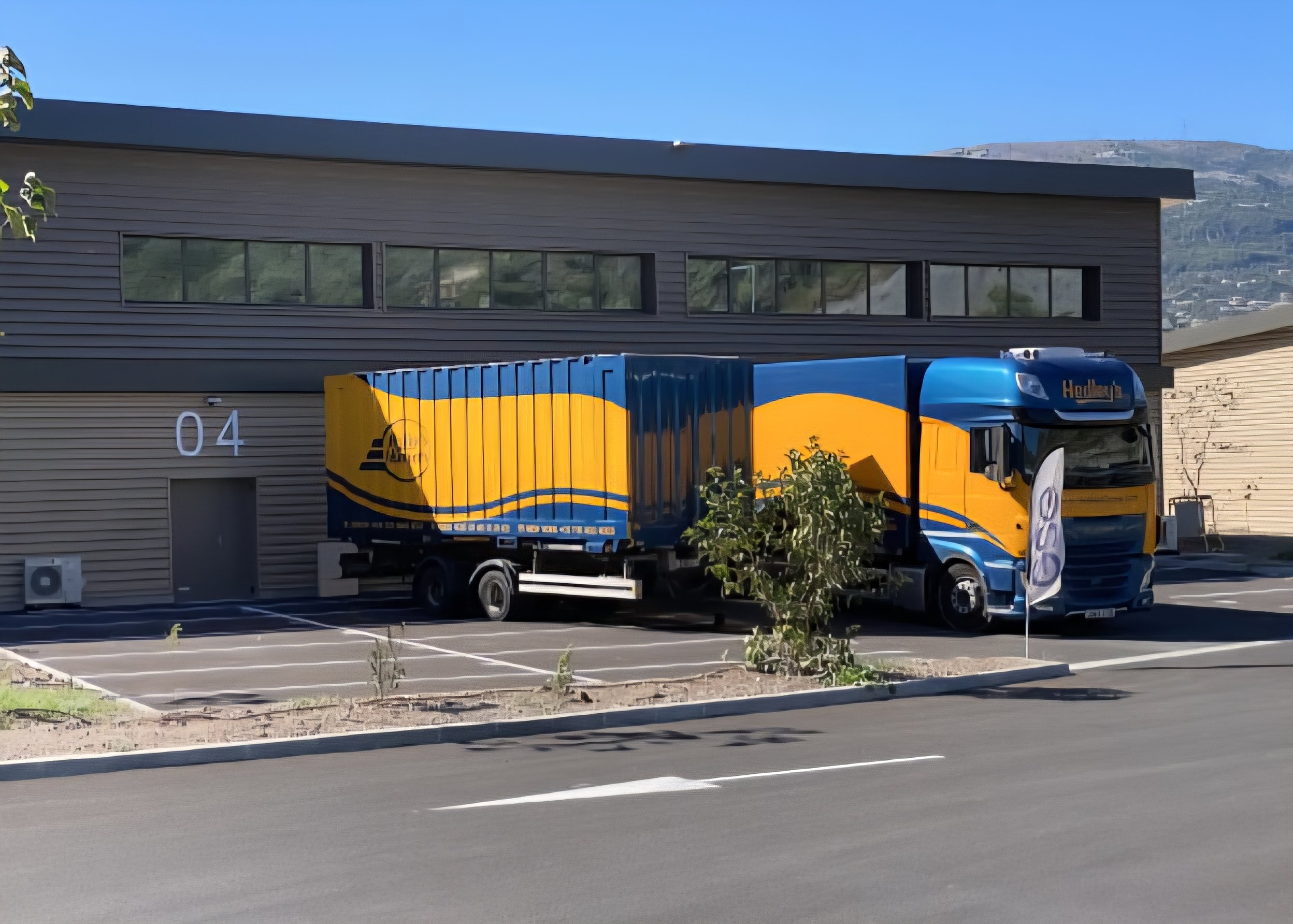 Camion de livraison coloré en bleu et jaune garé devant un bâtiment avec le numéro 04, sous un ciel clair.