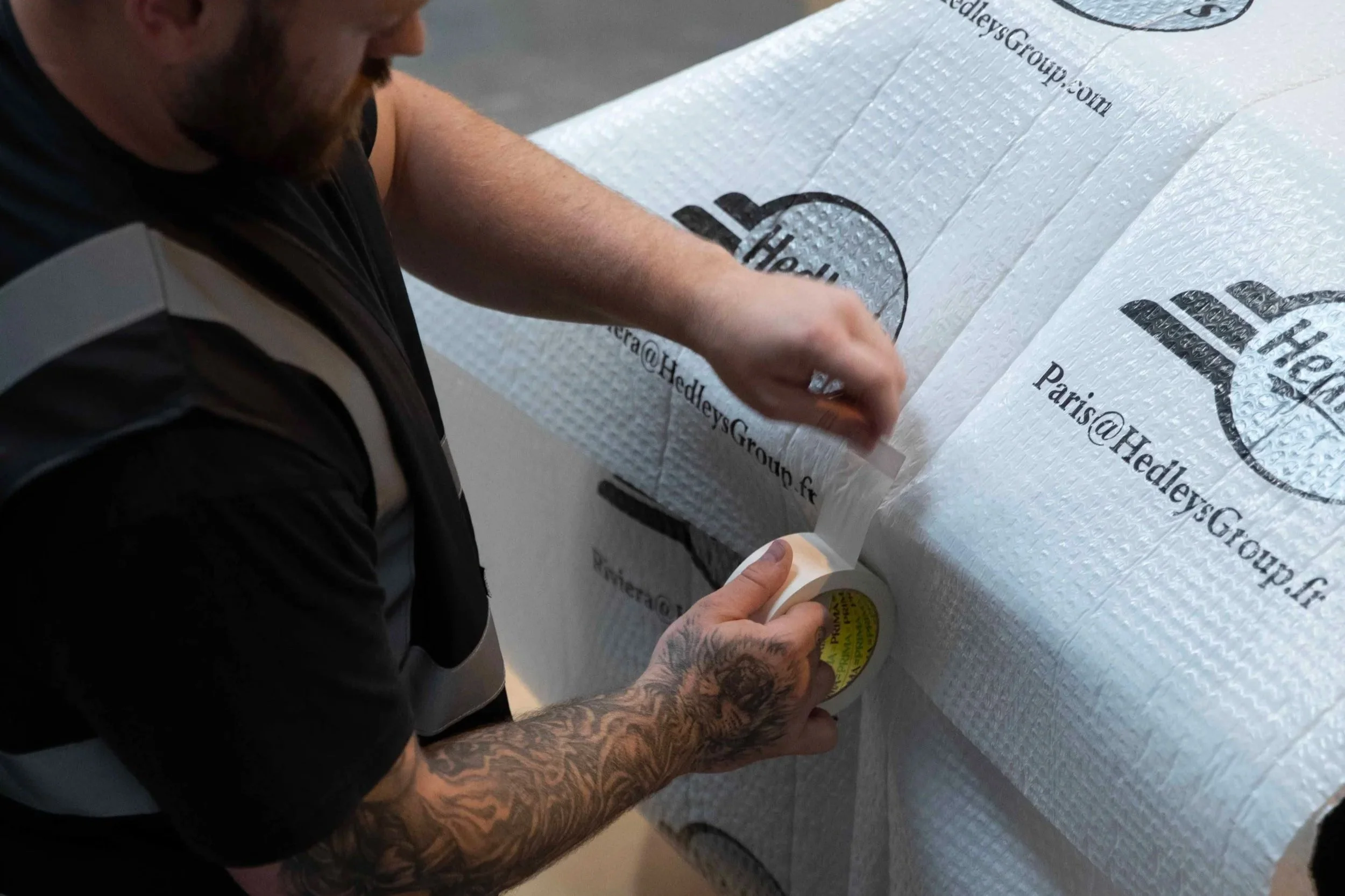 Une personne avec des tatouages appliquant du ruban adhésif sur une grande surface recouverte d'un matériau blanc, portant une veste noire avec une bande grise, lors d'une opération de collage ou d'emballage.