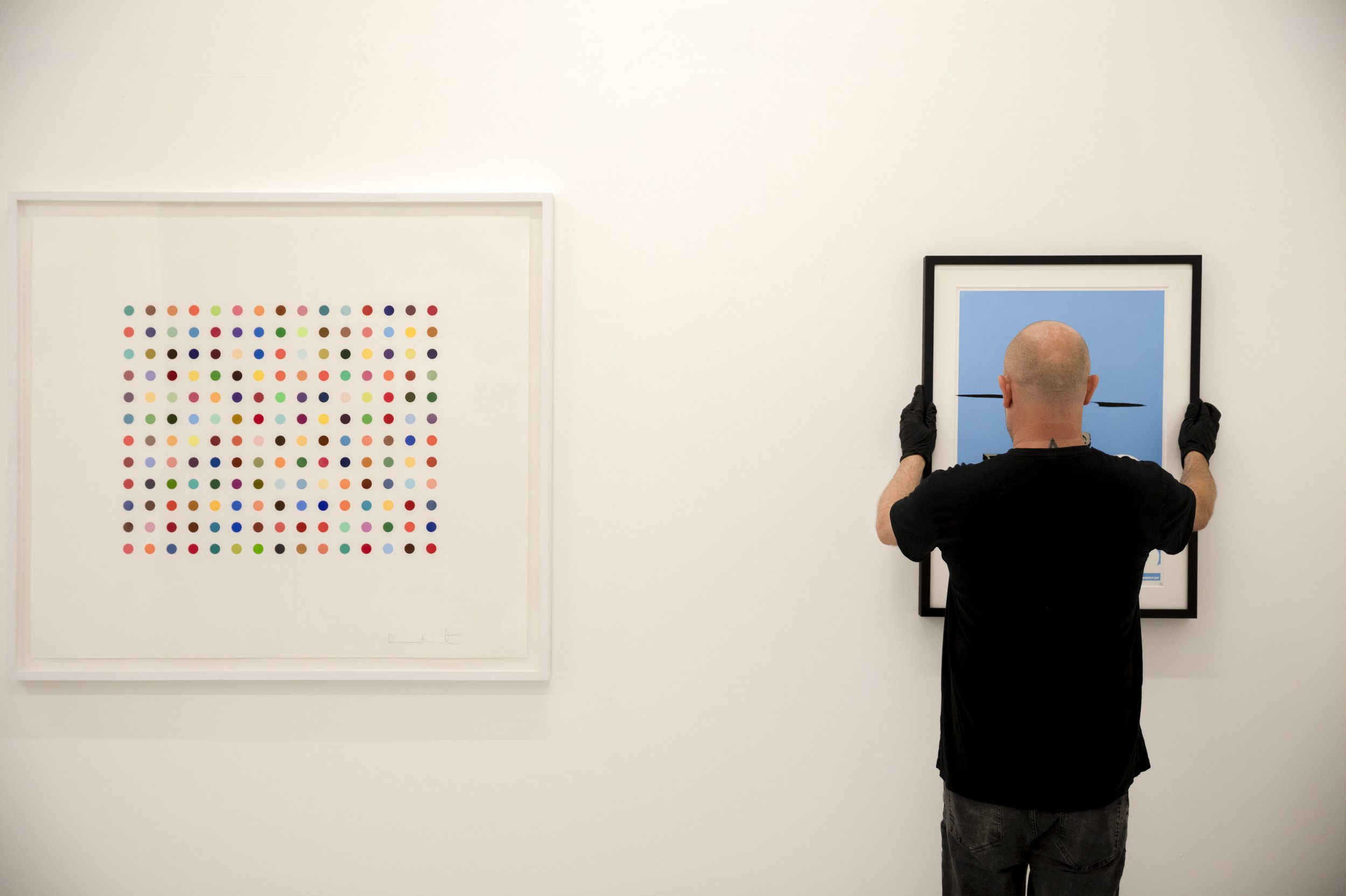 Un homme vu de dos, portant des gants noirs, accroche une œuvre d'art à un mur blanc. Deux œuvres d'art abstrait, l'une avec des points colorés et l'autre avec un fond bleu et des lignes noires, sont visibles.
