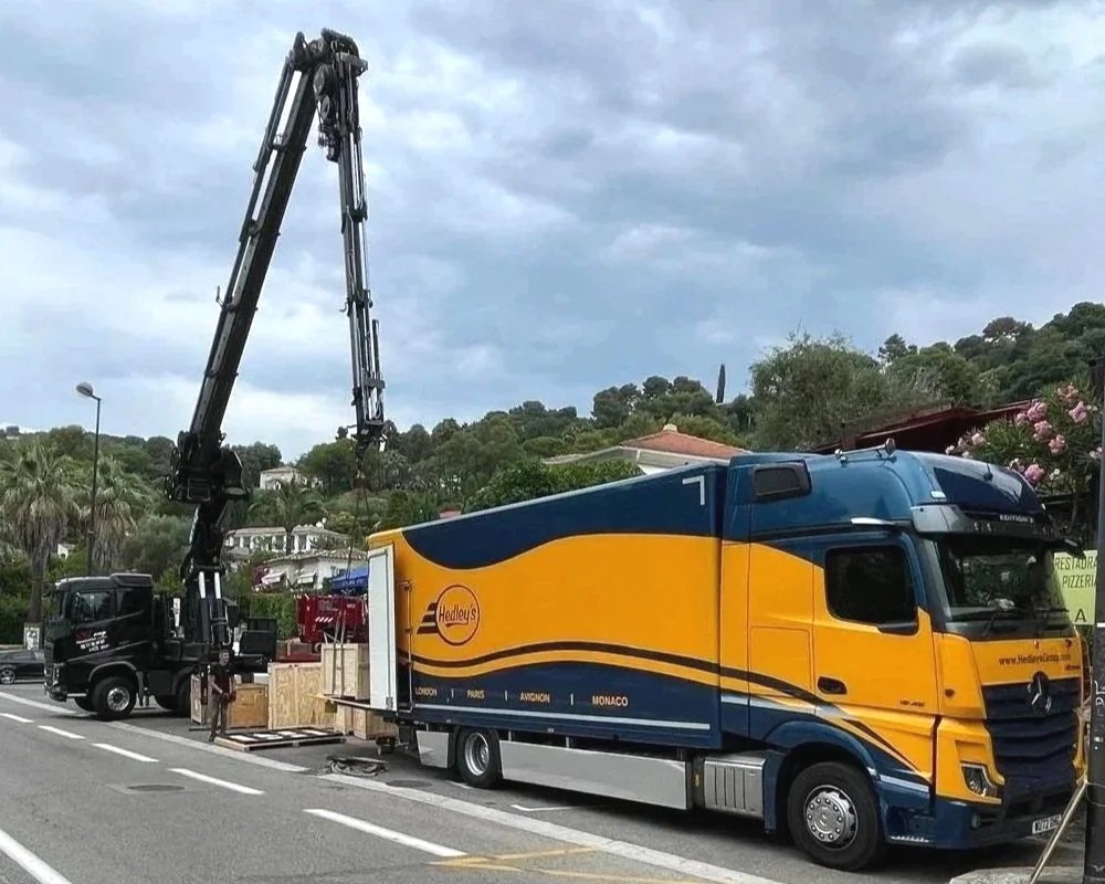 Un camion de déménagement avec une grue déchargé des meubles sur une route. Arbres et maisons en arrière-plan.