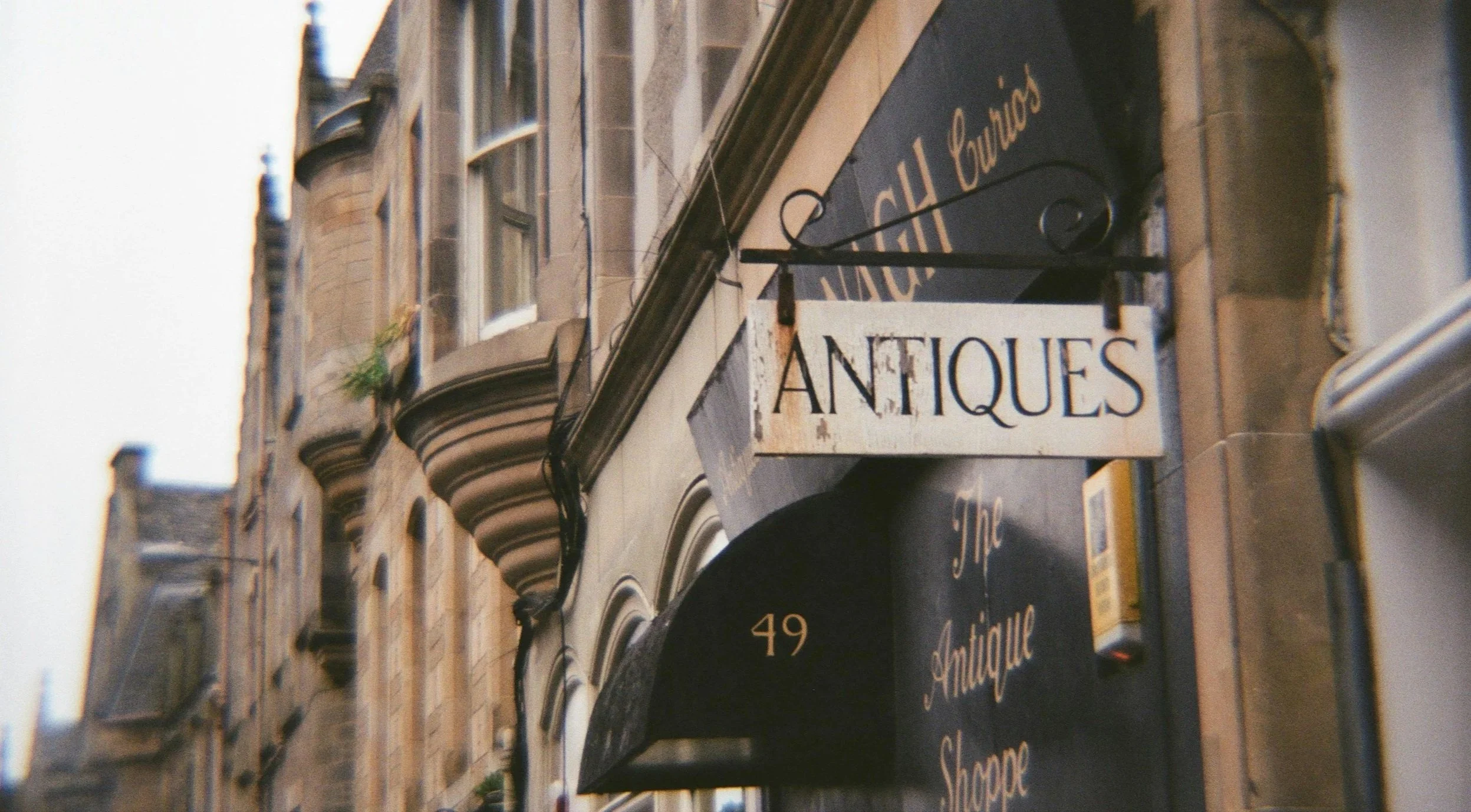 Une façade de boutique d'antiquités à l'angle d'une rue, avec un panneau en bois blanc portant le mot «ANTIQUE» suspendu par des crochets noirs. L'adresse »49« est visible sur un rideau noir à l'entrée.