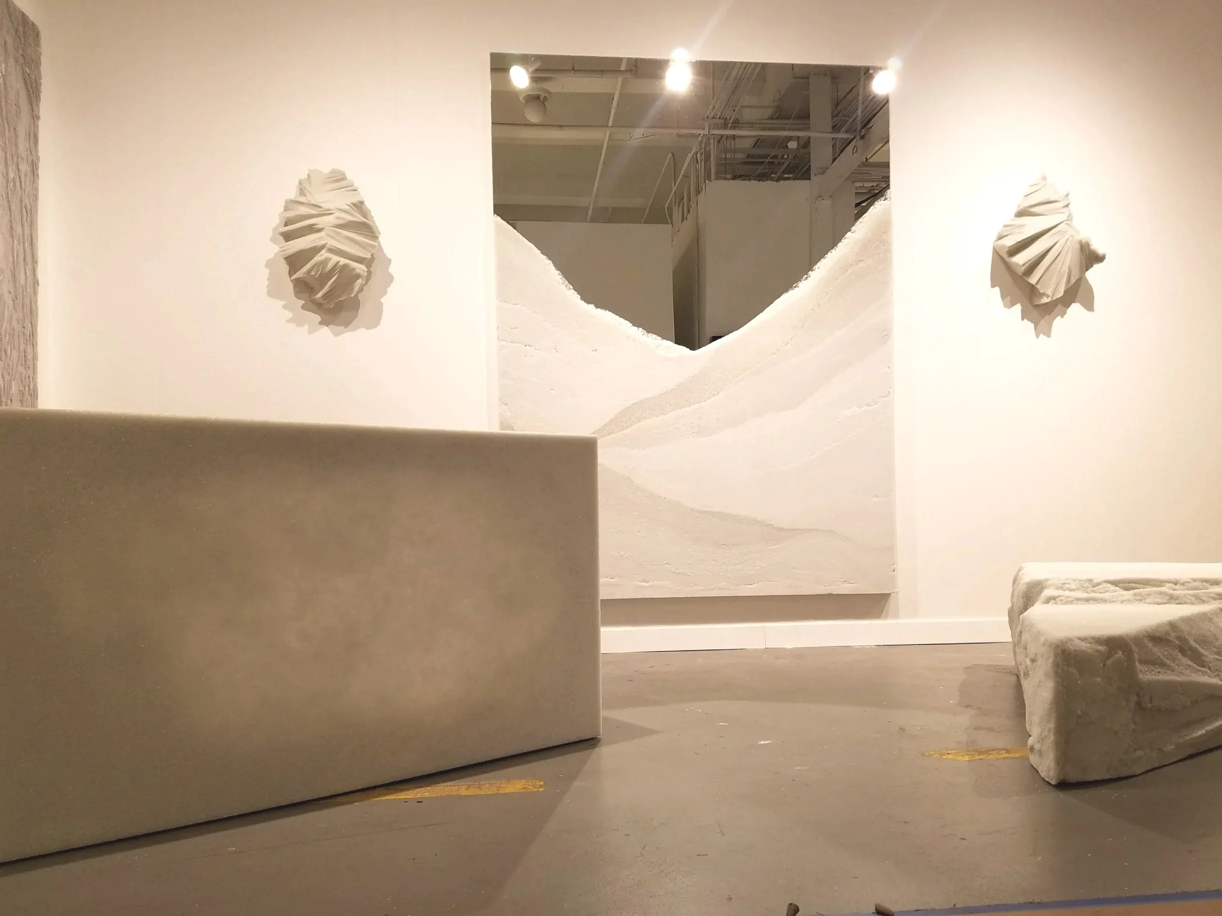 Installation artistique avec deux sculptures en papier sur un mur blanc, un tableau représentant une montagne blanche, et deux blocs de pierre posés au sol.