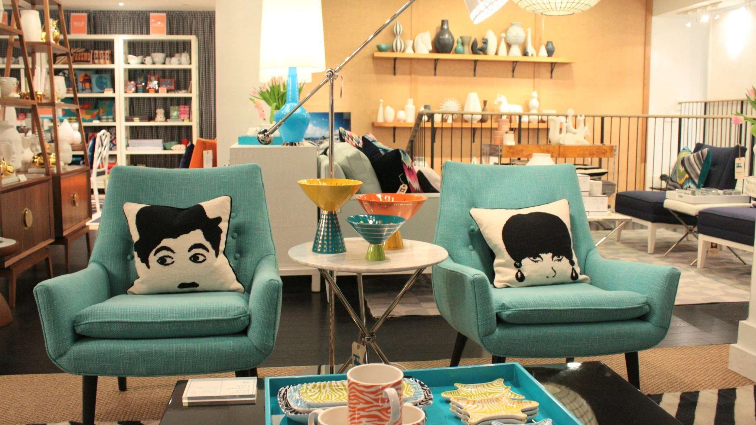 Une boutique de décoration intérieure avec deux fauteuils turquoise, des coussins avec des visages, une petite table avec des céramiques colorées, et des étagères avec des objets décoratifs blancs et noirs.