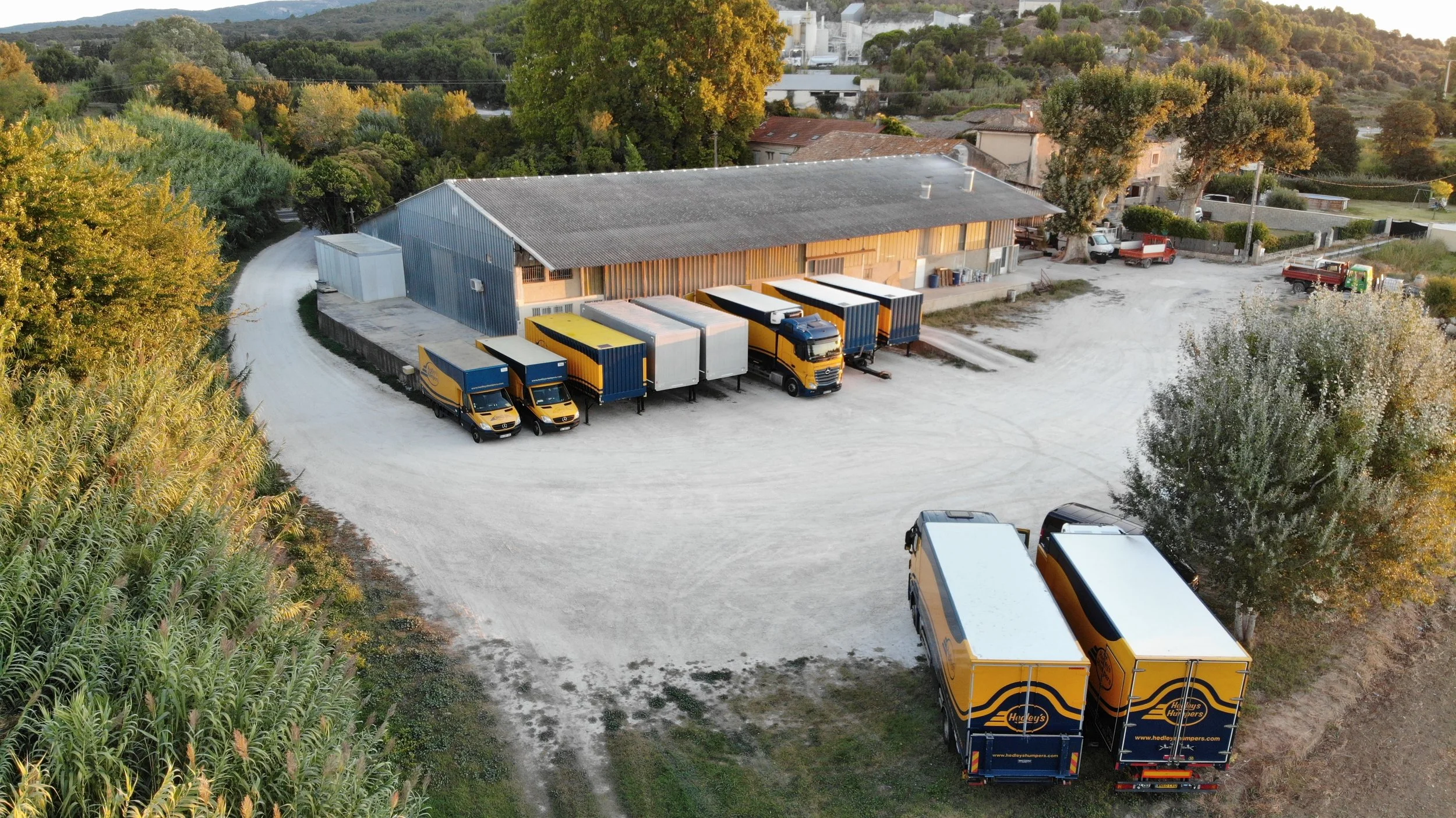 Vue aérienne d'un entrepôt avec plusieurs camions de livraison stationnés devant, entouré de végétation et de maisons dans une zone rurale.