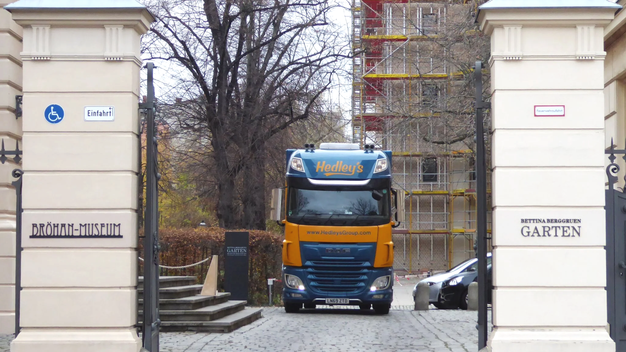 A blue and yellow Hedley's delivery truck parked in front of an entrance gate to a garden at the Bröhan Museum. The gate has signs indicating wheelchair accessibility and fire department access, with a construction scaffold visible behind the gate.