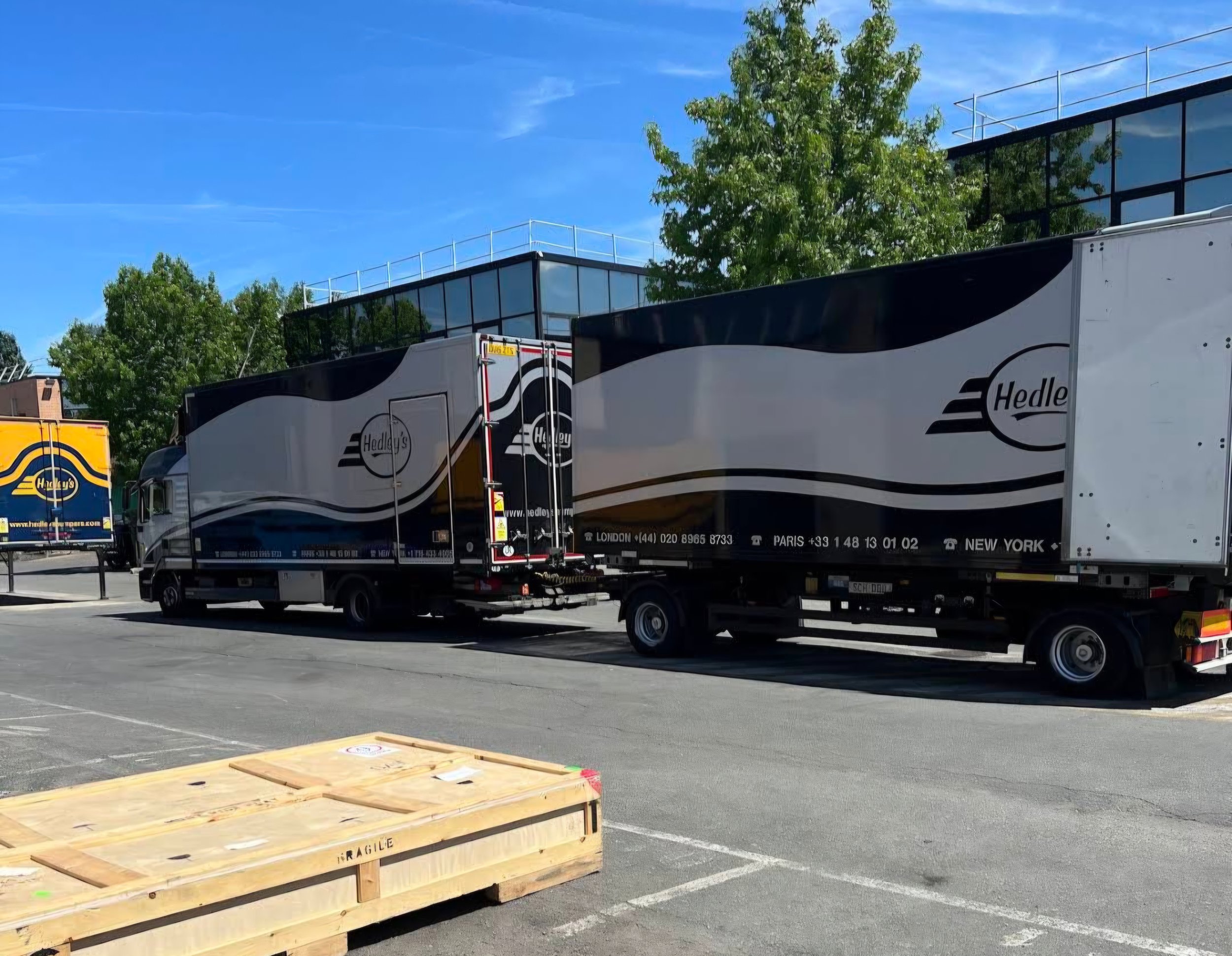 Deux camions de livraison Hedley's stationnés dans un parking, un bâtiment moderne avec des fenêtres en arrière-plan, un ciel bleu avec quelques nuages, et un conteneur en bois marqué 'Fragile' au premier plan.