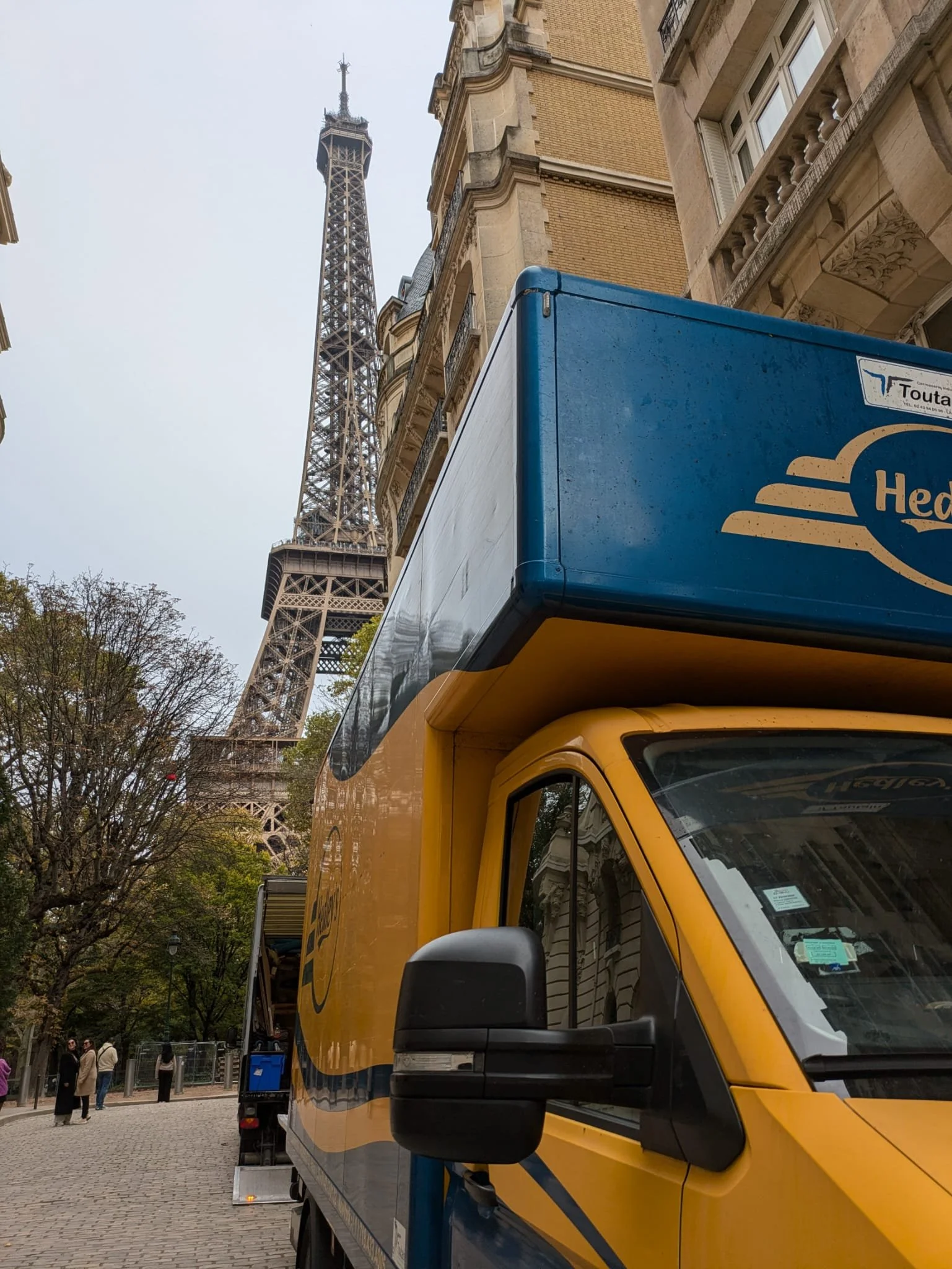 looking up at the Eiffel Tower in Paris, France, with a yellow and blue delivery truck in the foreground and some people walking on a cobblestone street