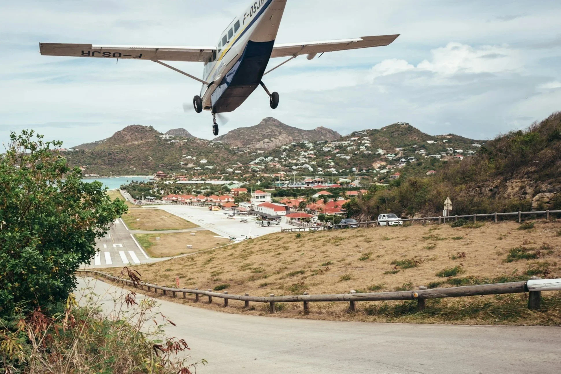 Petite avion en vol au-dessus d'un aéroport avec un paysage montagneux et résidentiel en arrière-plan.