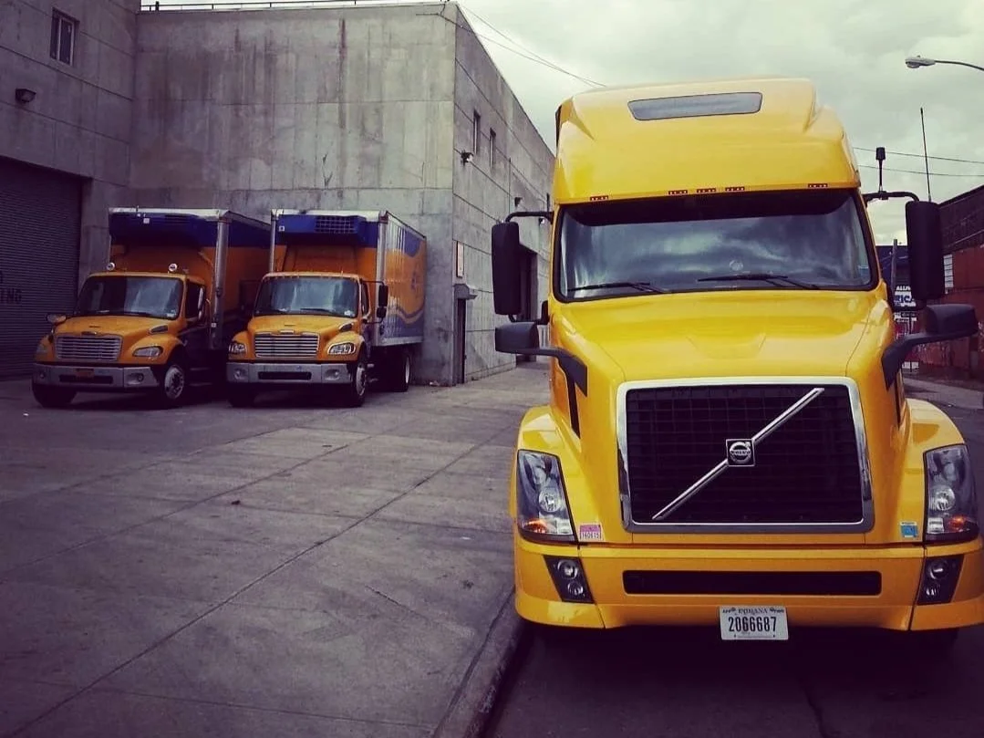Four yellow delivery trucks parked on a concrete sidewalk near a building, with three trucks lined up against the wall and one in the foreground facing forward.