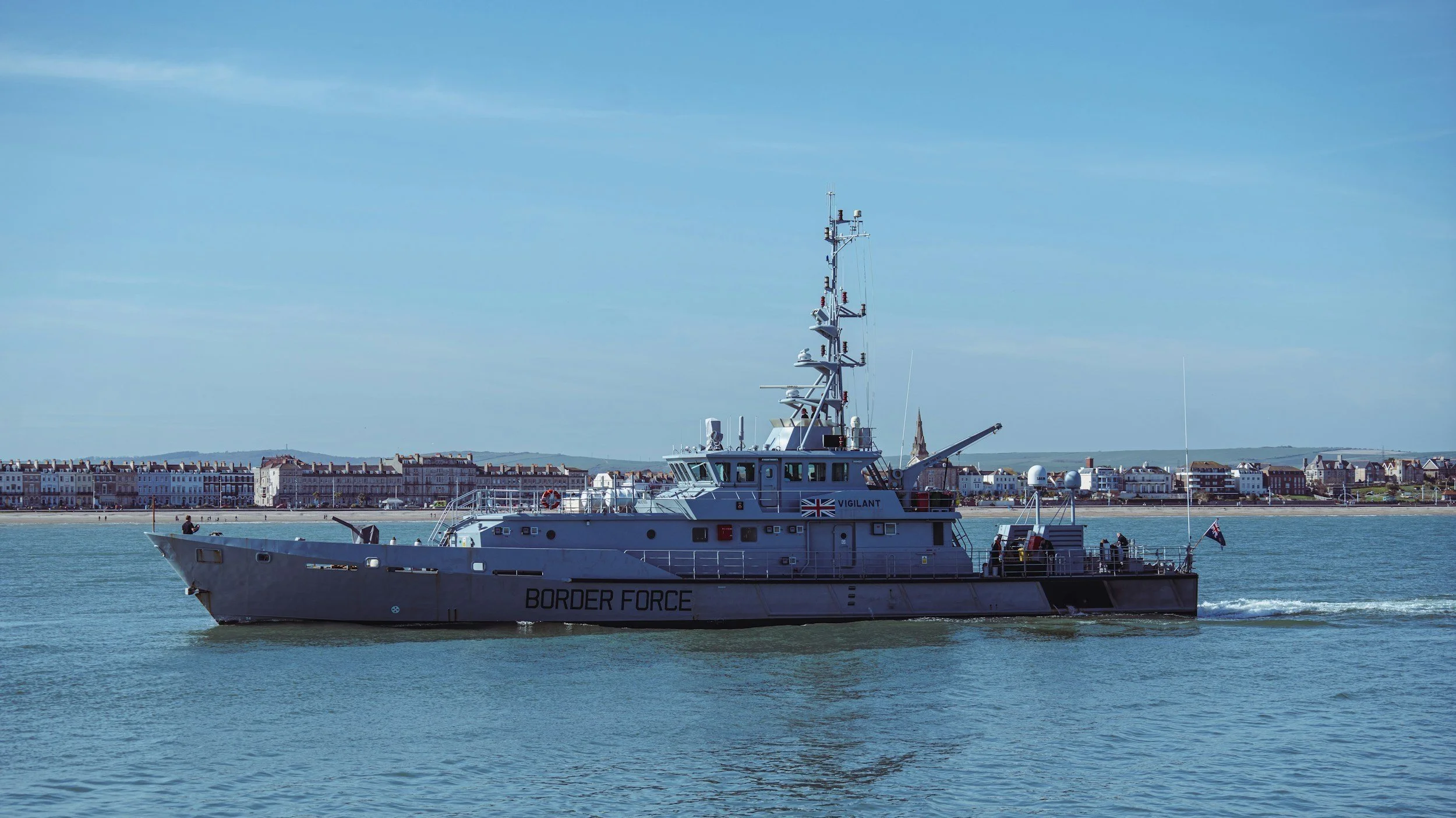 Bateau des douanes de la garde frontière naviguant sur l'eau avec un fond de ville côtière et un ciel bleu.