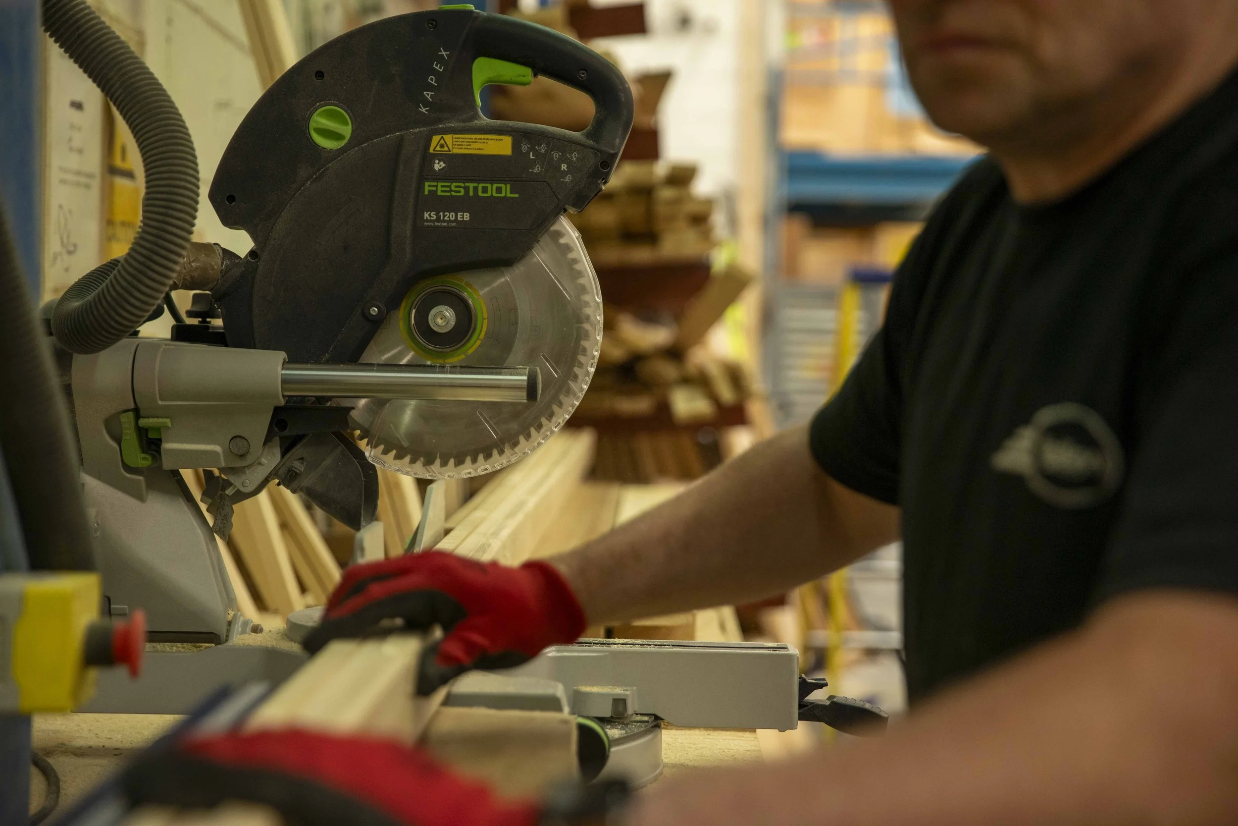 A man wearing a black shirt and red gloves operating a miter saw in a woodworking shop, cutting a piece of wood.