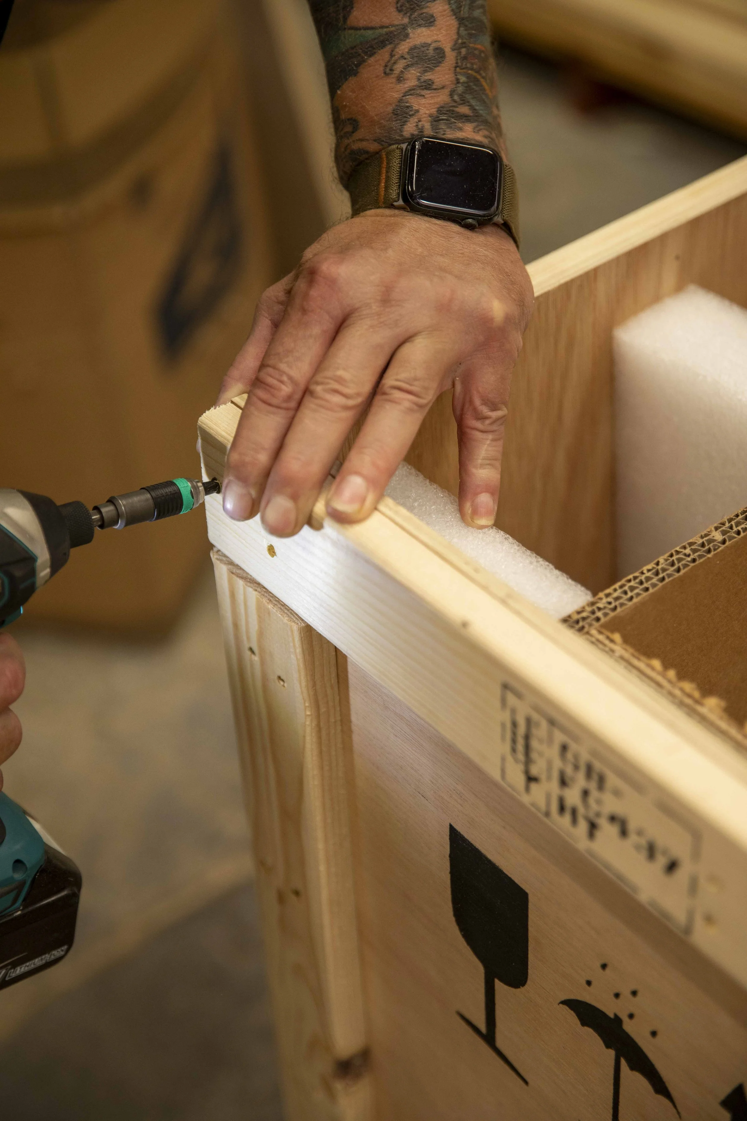 A person using a drill to assemble a wooden crate or box, with foam padding and packing material inside.