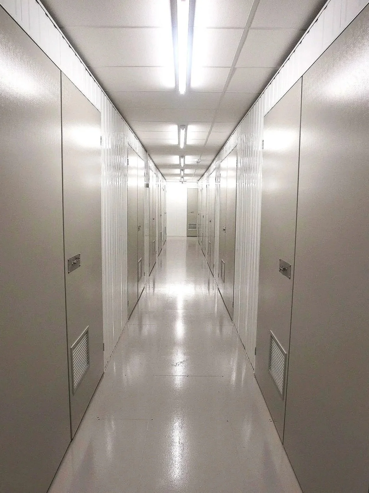 Empty, well-lit hallway with lockers on both sides, smooth white floor, ceiling lights, and a door at the end.