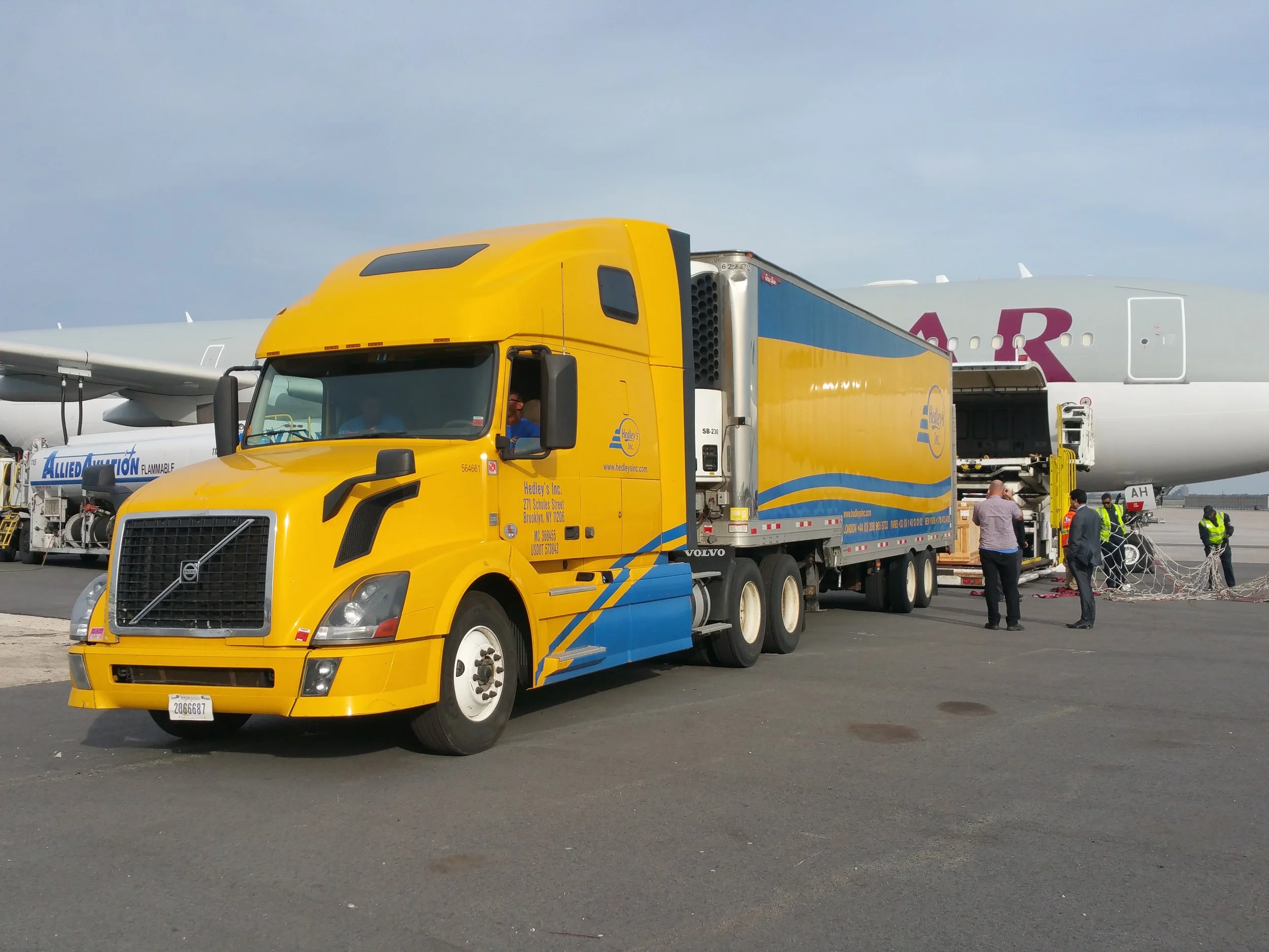 Un camion de transport de bagages jaune et bleu stationne devant un grand avion dans un aéroport. Des employés et des personnes regardent ou travaillent près de l'avion, qui est en cours de chargement ou de déchargement.