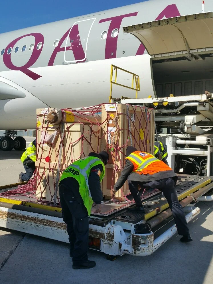 Airport cargo workers unload a pallet of goods from an airplane using a conveyor belt, with a large cargo container on the tarmac and workers in safety vests handling the cargo.
