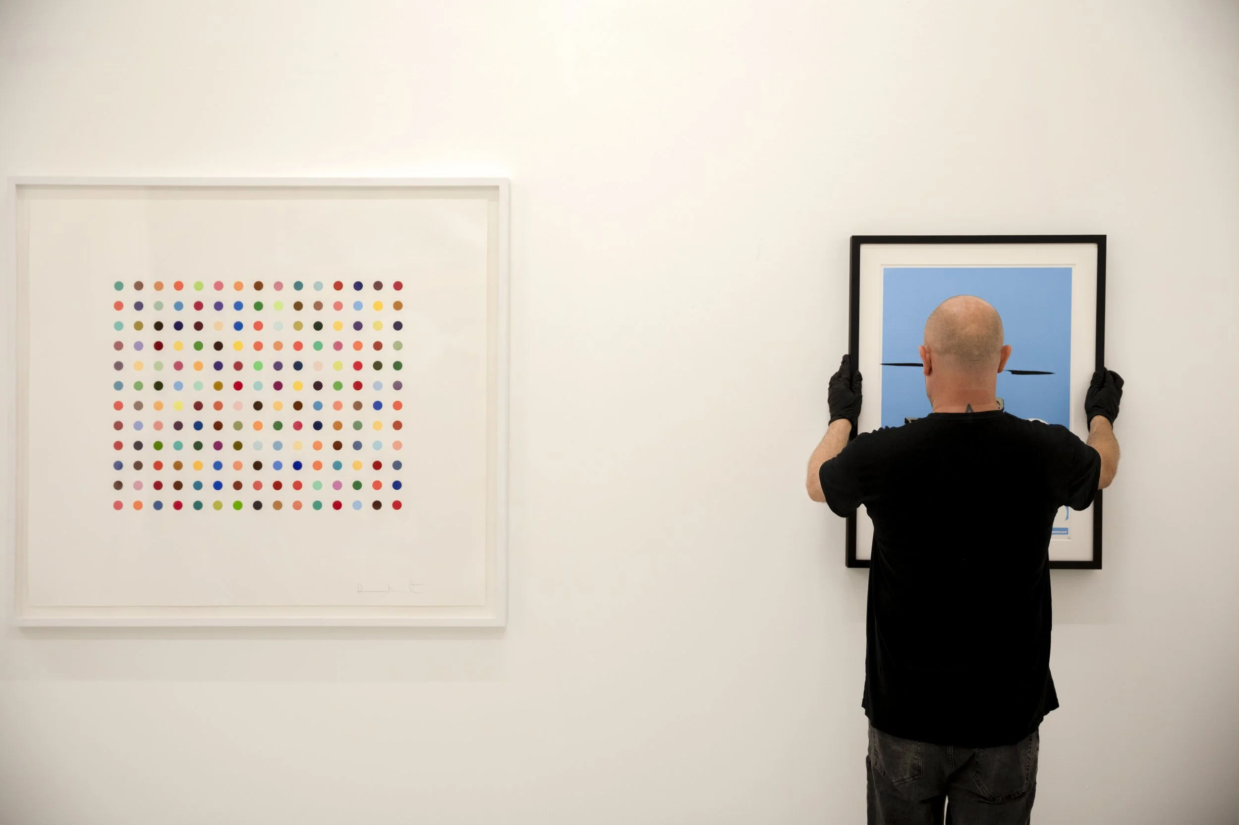 Un homme sans cheveux, portant des gants noirs, accroche une œuvre d'art dans une galerie. La pièce à gauche est une affiche colorée avec un motif de points de différentes couleurs sur fond blanc. La pièce à droite est une image bleue avec deux lignes horizontales et une verticale.