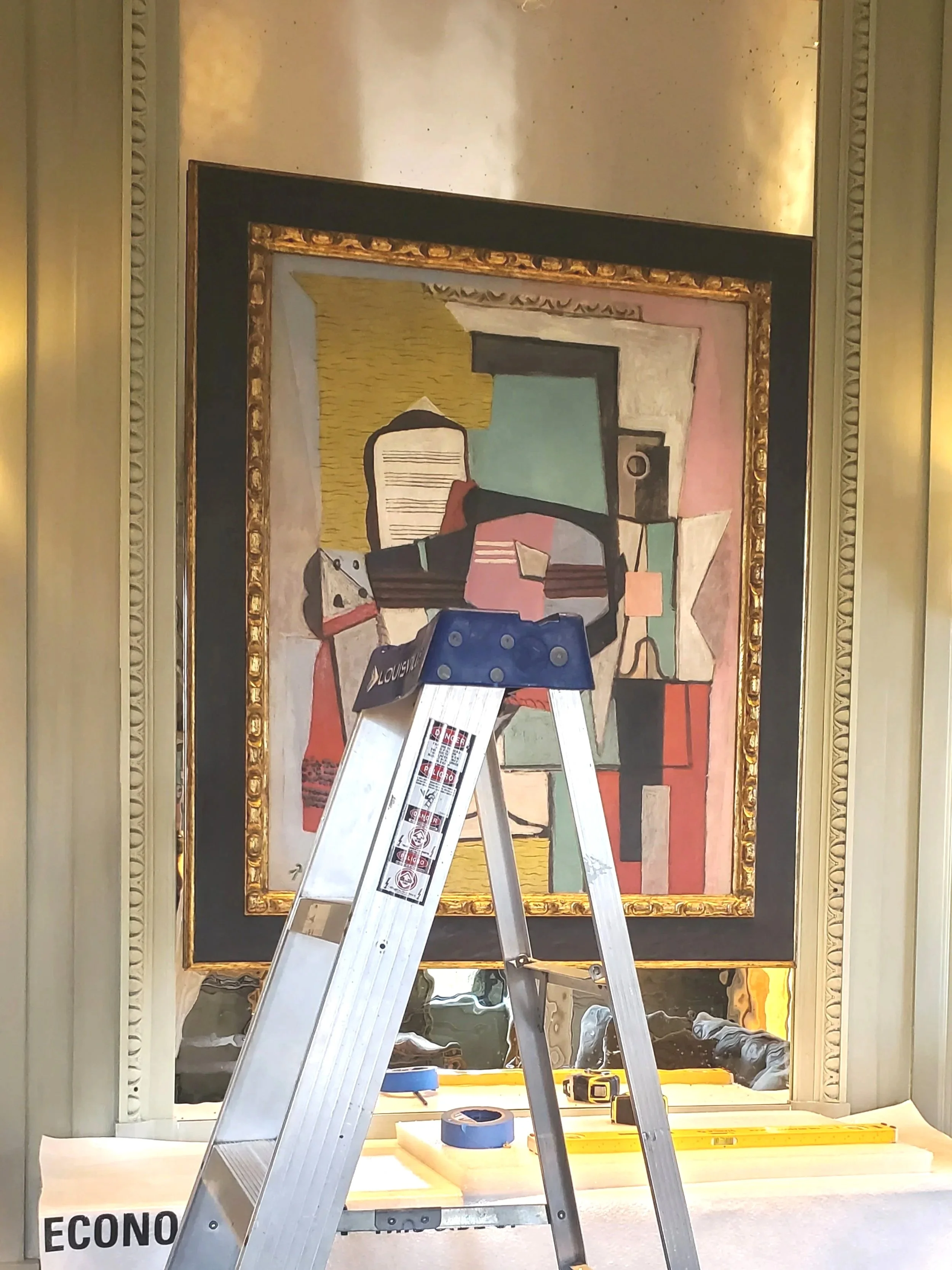 A framed abstract painting on a wall behind a silver step ladder inside a room. The painting features geometric shapes and bold colors, and the wall has decorative molding.