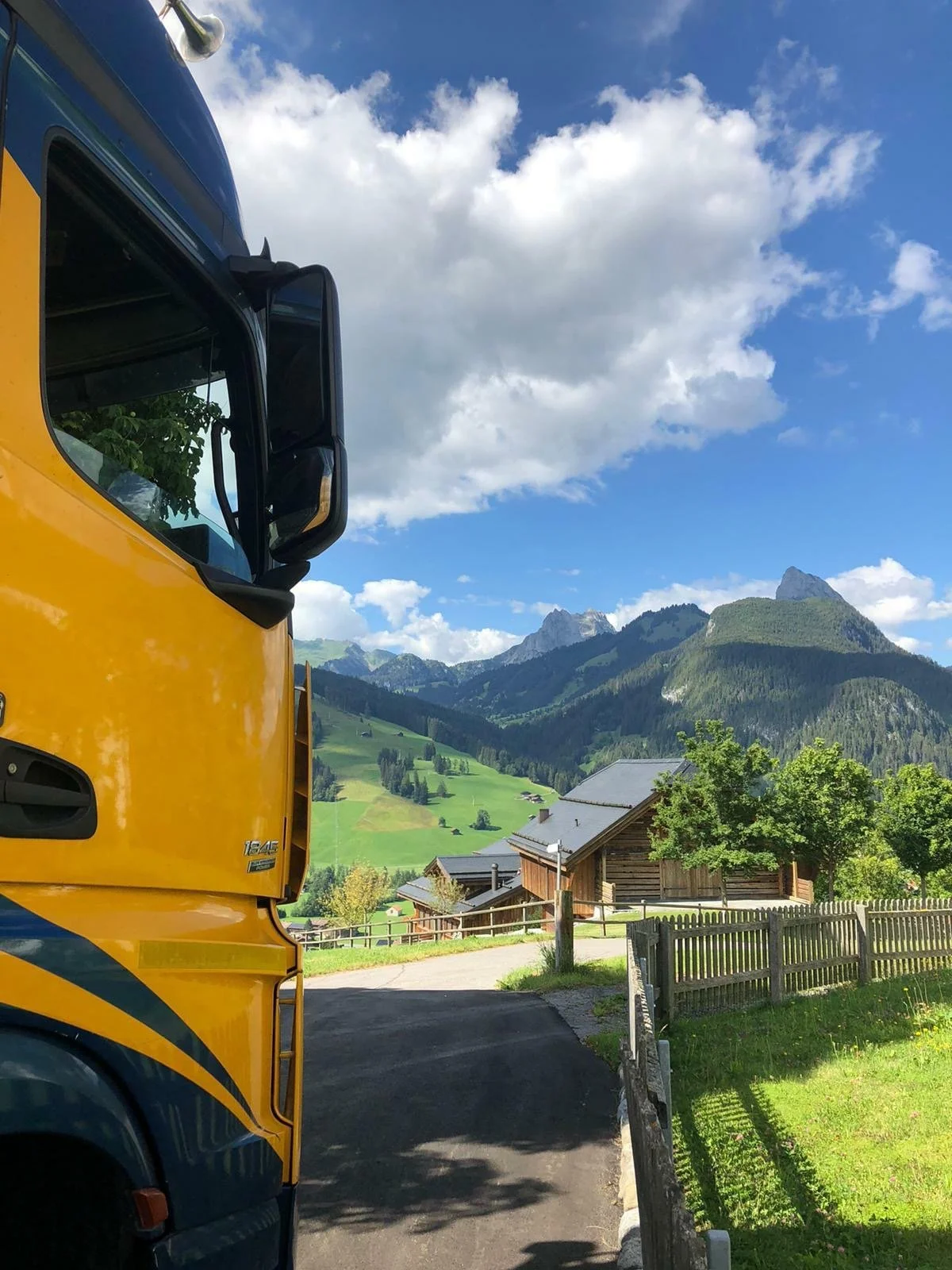 A yellow and blue bus parked on a road in a rural area with green hills, trees, wooden houses, and mountains in the background under a partly cloudy sky.